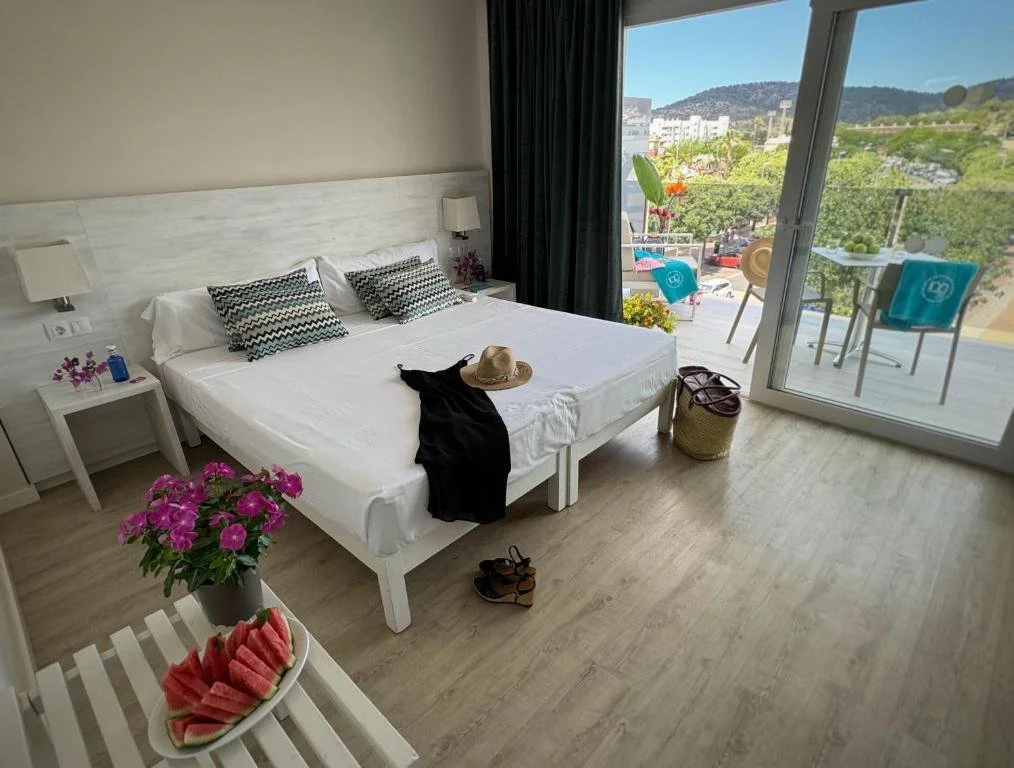 Hotel room with a large bed, two side tables, a balcony with outdoor seating, and a view of green hills and buildings outside.