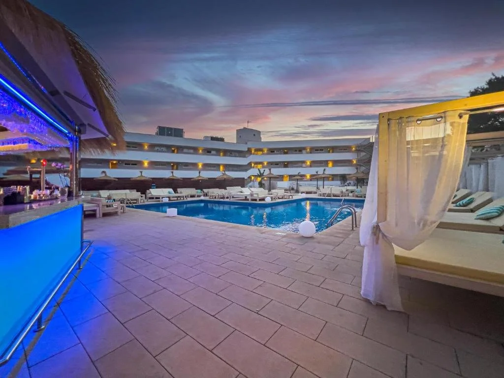 Swimming pool area at a resort during sunset with lounge chairs, cabanas, and colorful sky.