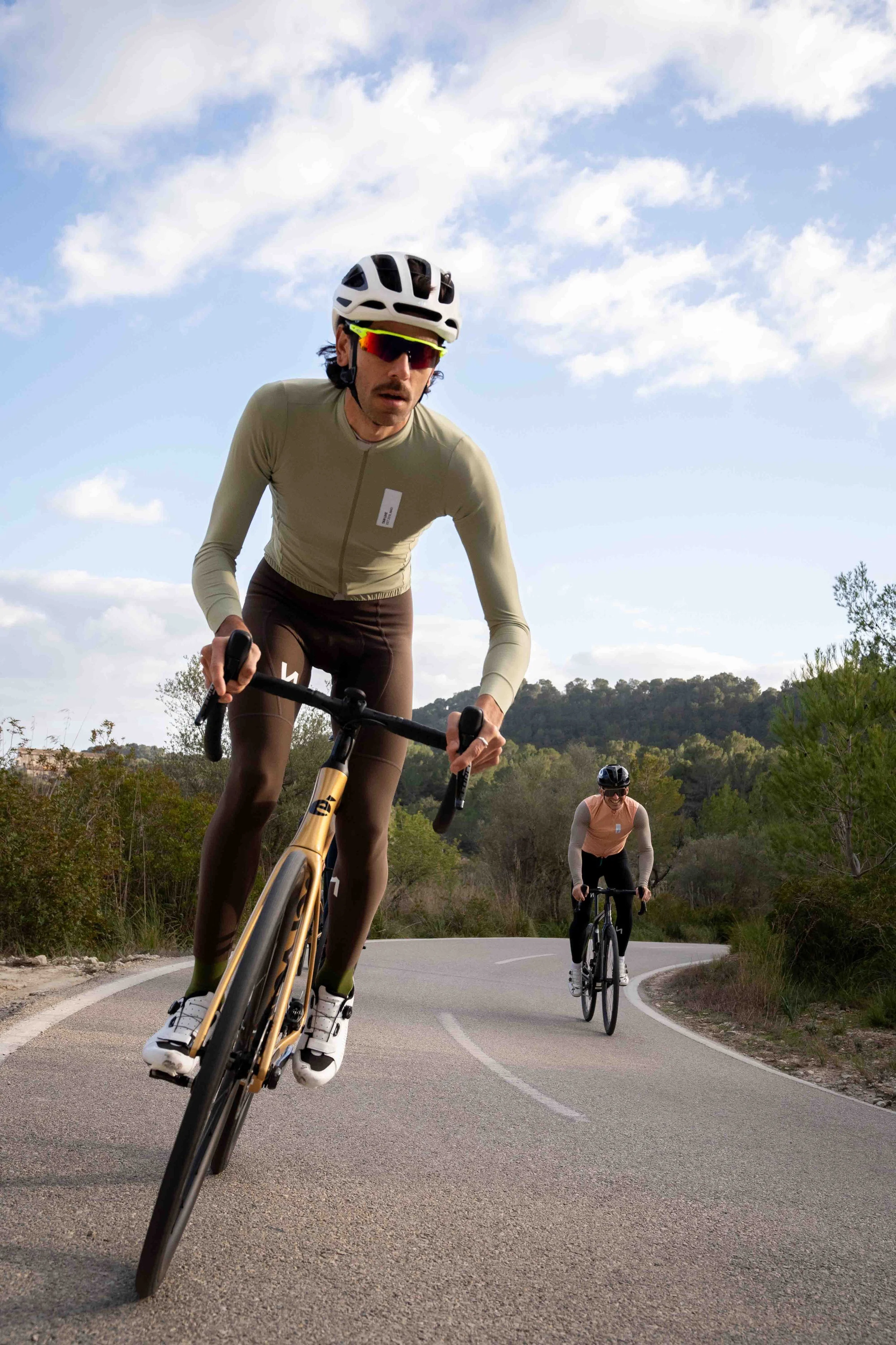 Two cyclists riding on a winding road in a rural area with trees and a partly cloudy sky.