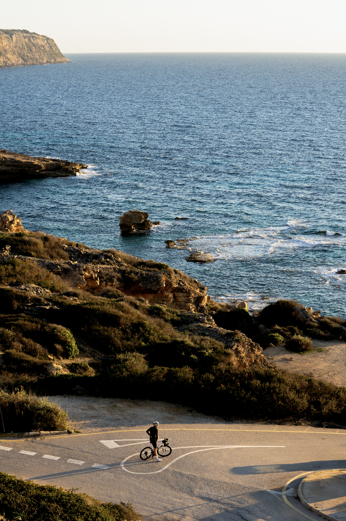 A person in cycling gear standing next to a bike on a winding road near the coast, with rocky cliffs and the ocean in the background during sunset.