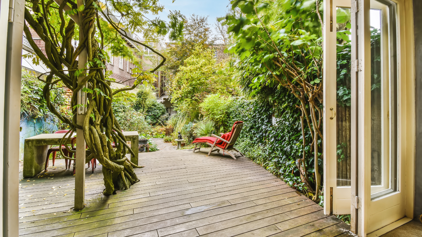 View of a lush garden patio with wooden deck, green plants, and trees. There is red outdoor furniture including a lounge chair with a shade on it, and a bench. The photo is taken from inside a house through an open door.