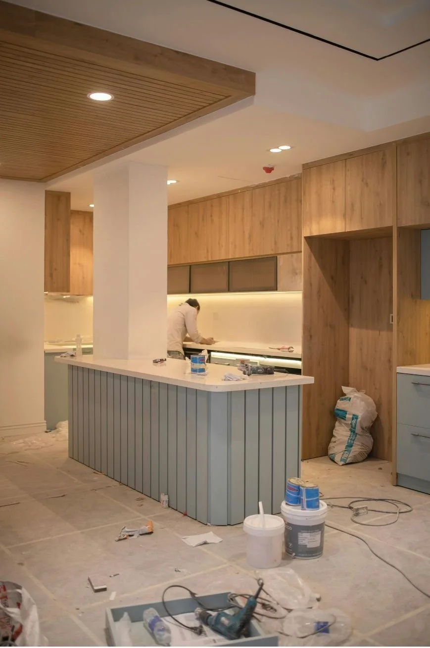 A person working in a kitchen under construction, with wood-paneled cabinets, a countertop, and construction tools and materials scattered on the floor.
