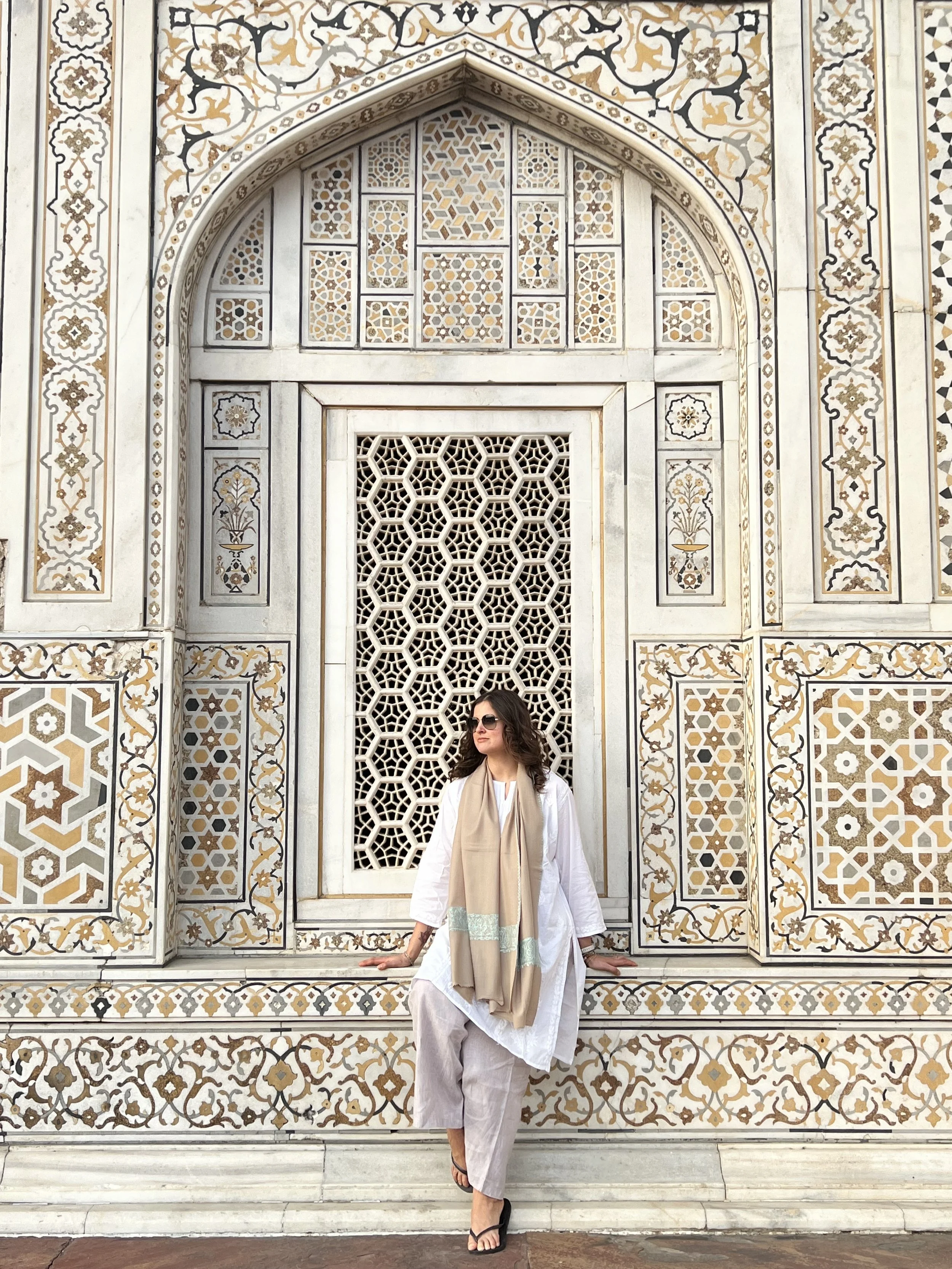 A woman in white traditional attire with a beige scarf, wearing sunglasses, leaning against an ornate white stone wall with intricate geometric and floral designs, in front of a lattice window and decorative archway.