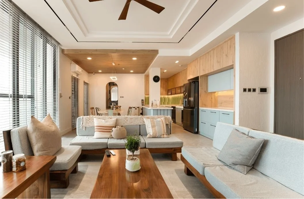 A modern, open-concept living room and kitchen with neutral tones, wooden accents, large windows with blinds, and contemporary furniture and decor.