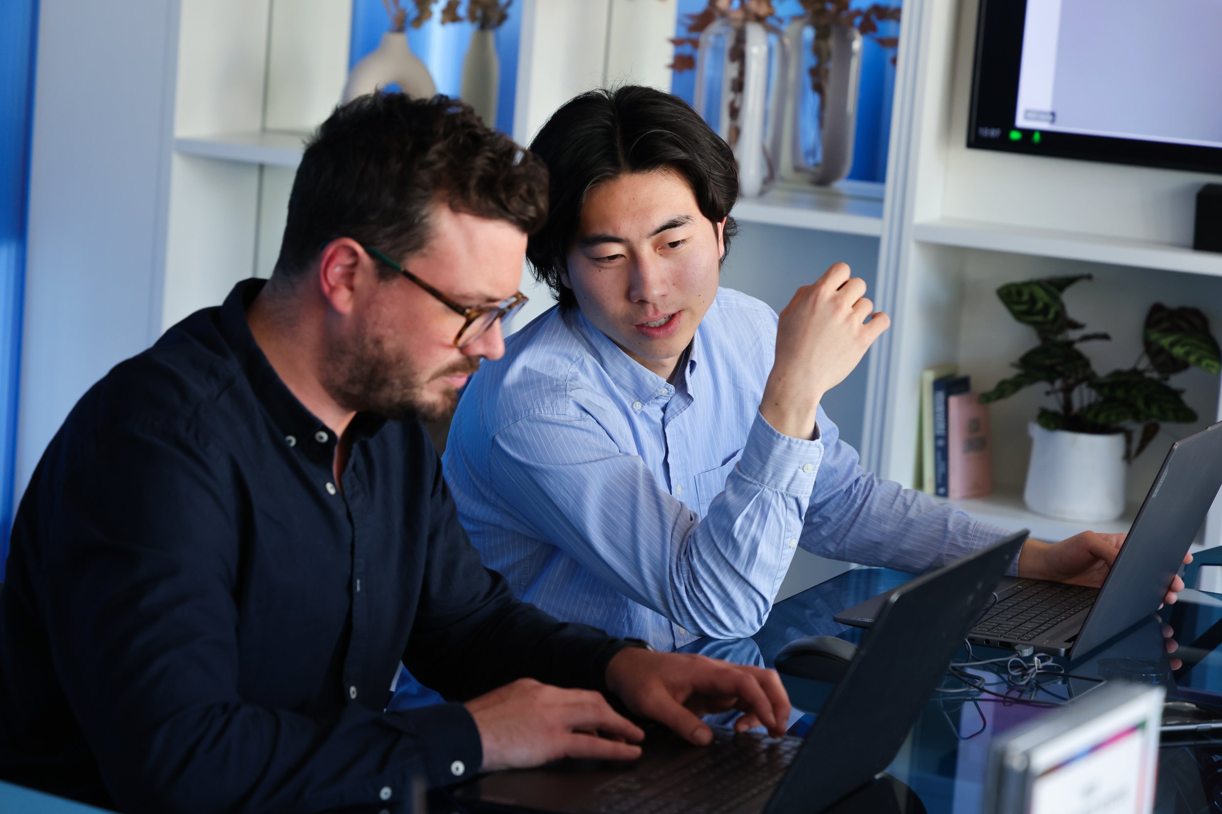 Two men working on laptops in an office, one in glasses and a dark shirt, the other with dark hair and a light shirt, engaged in a discussion.