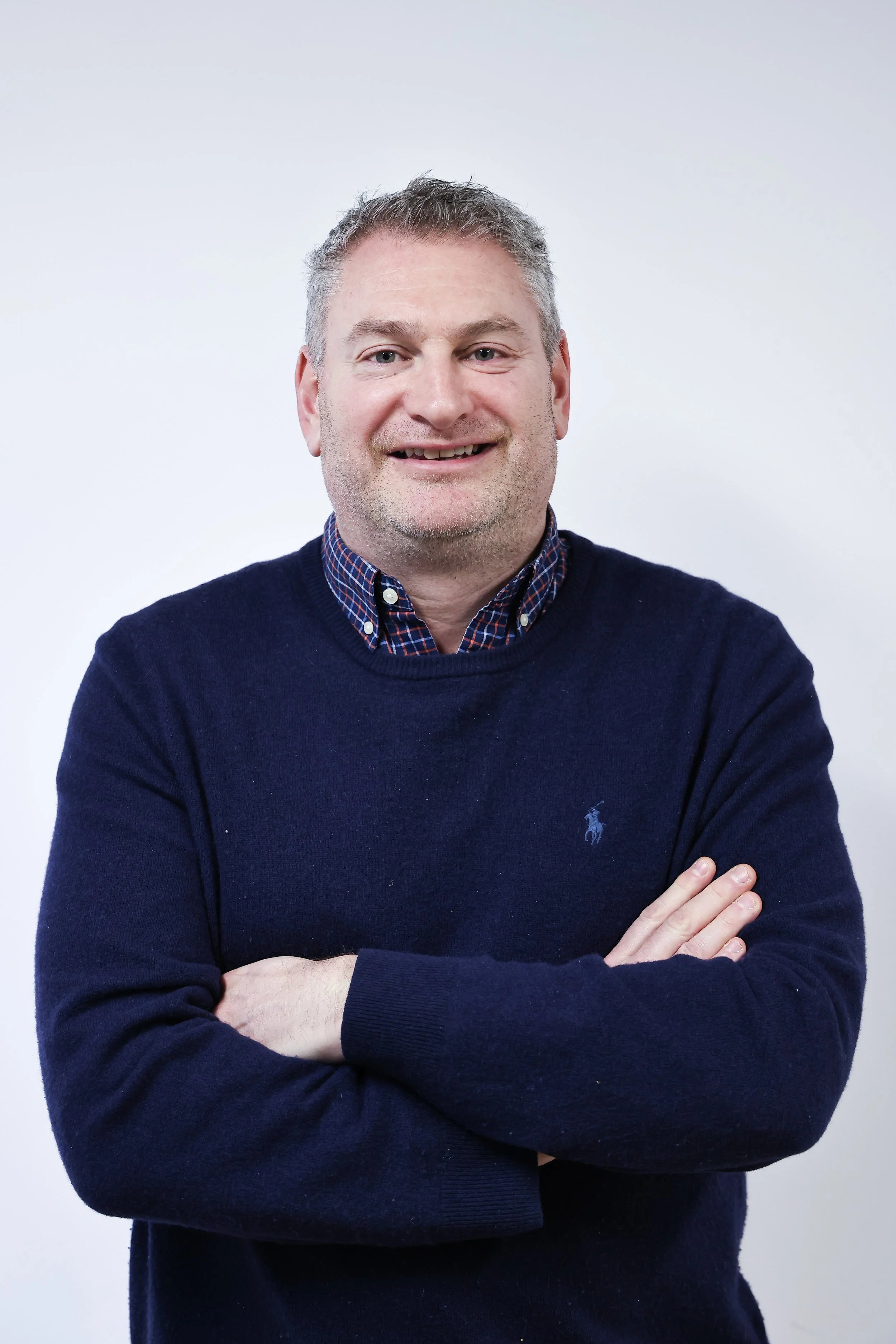 A middle-aged man with short gray hair and blue eyes smiling, wearing a black sweater with a small Polo logo and a collared shirt underneath, standing against a light gray background.