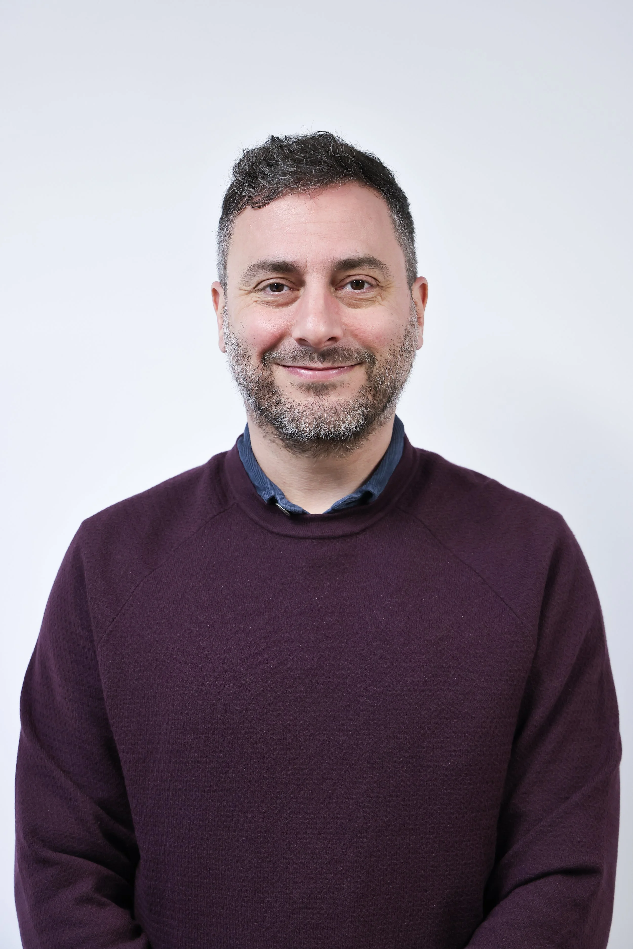 A smiling man with short dark hair and a beard, wearing a dark button-up shirt, standing against a light gray background.