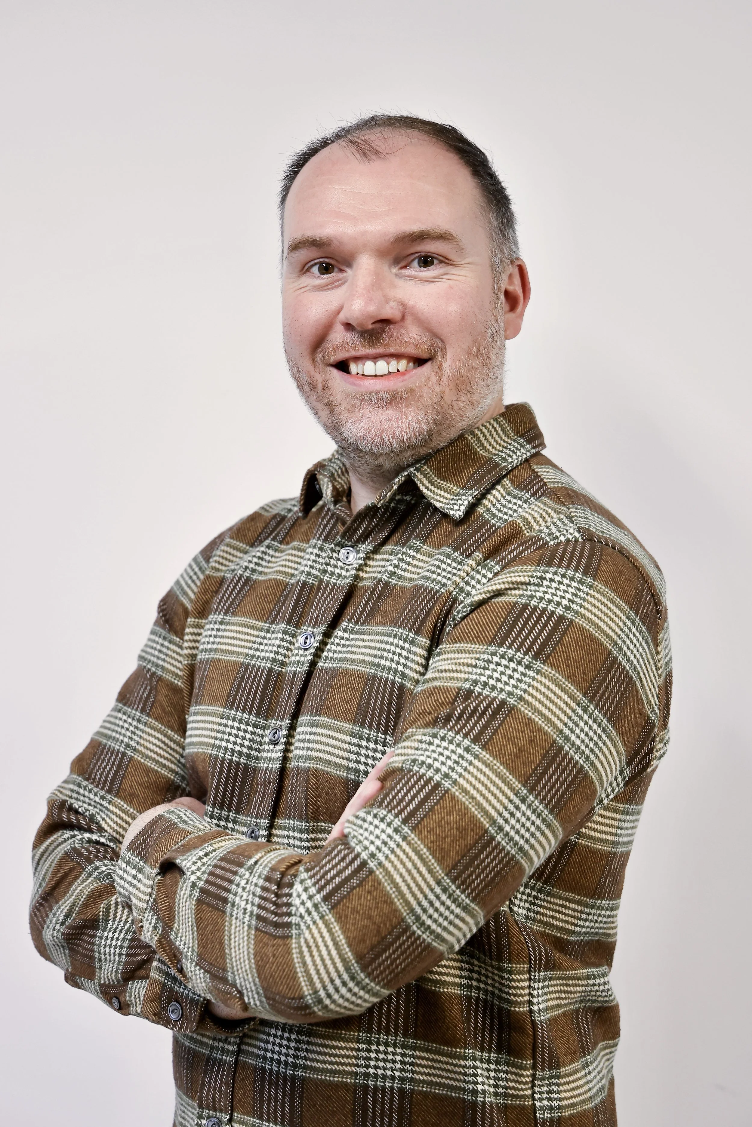 A smiling man with light brown hair and a beard, wearing a blue button-up shirt, standing against a plain gray background.