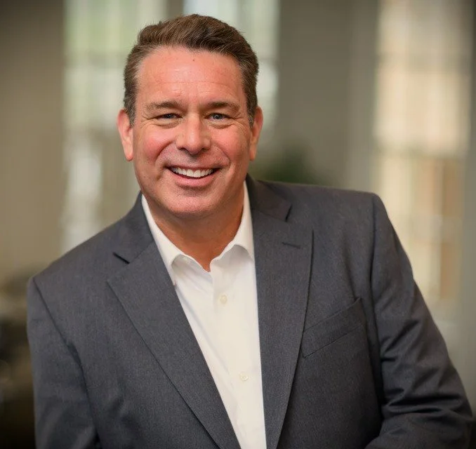 A smiling man in a gray suit jacket and white dress shirt looks directly at the camera in a professional setting.