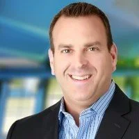 A smiling man in a dark blazer and blue shirt standing in an office or corporate environment.
