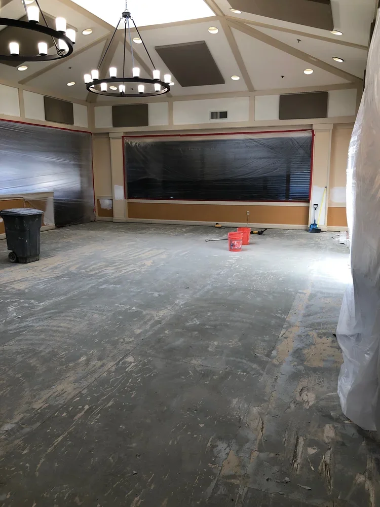 An empty room undergoing renovation with a partially stripped floor, plastic covering on the walls and windows, a trash can, a broom, and two orange buckets.