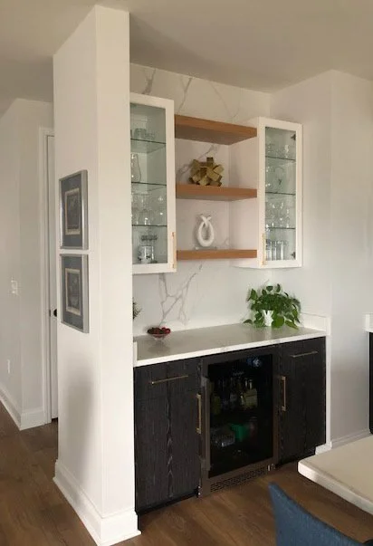 A built-in cabinet with glass shelves, decorated with vases and ornaments, mounted on a white wall with a marble-patterned backsplash. Below the cabinet is a countertop with a potted plant and a small bowl, underneath which is a mini-fridge with blac