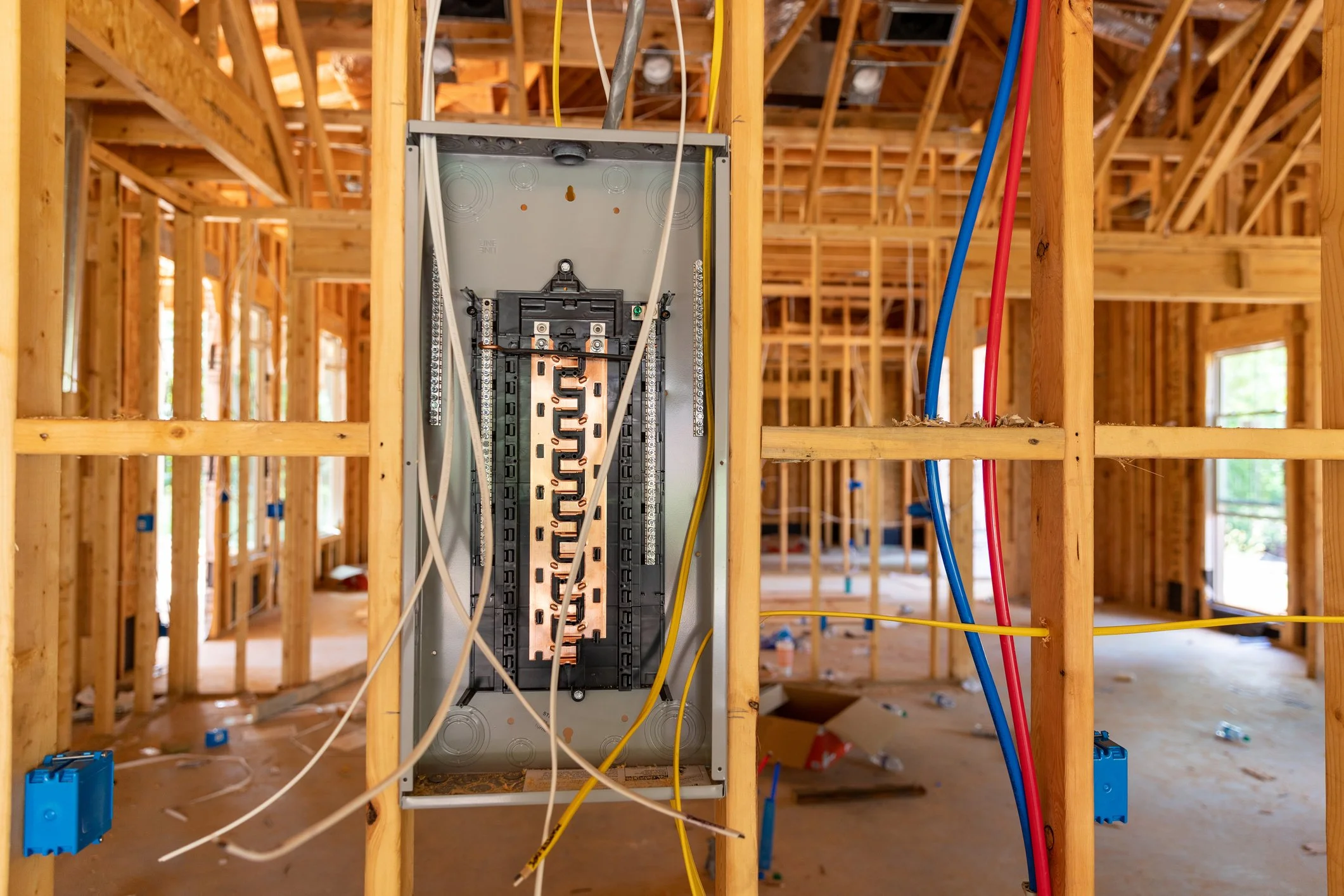 An electrical panel is installed in the wooden framing of a house under construction, with red, blue, yellow wires connected to it.