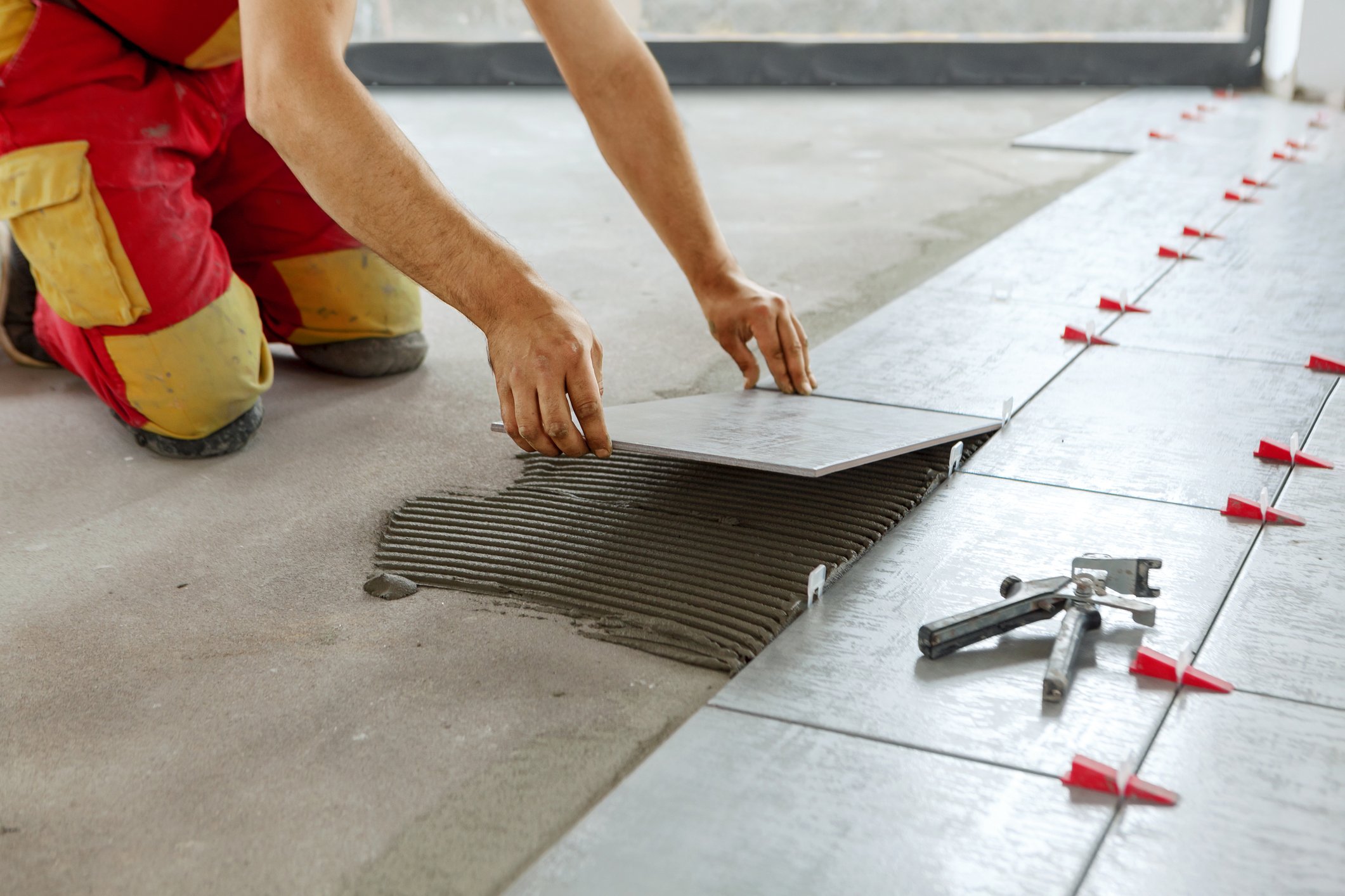 A worker installing tiles on a floor, kneeling and using a notched trowel to spread adhesive.