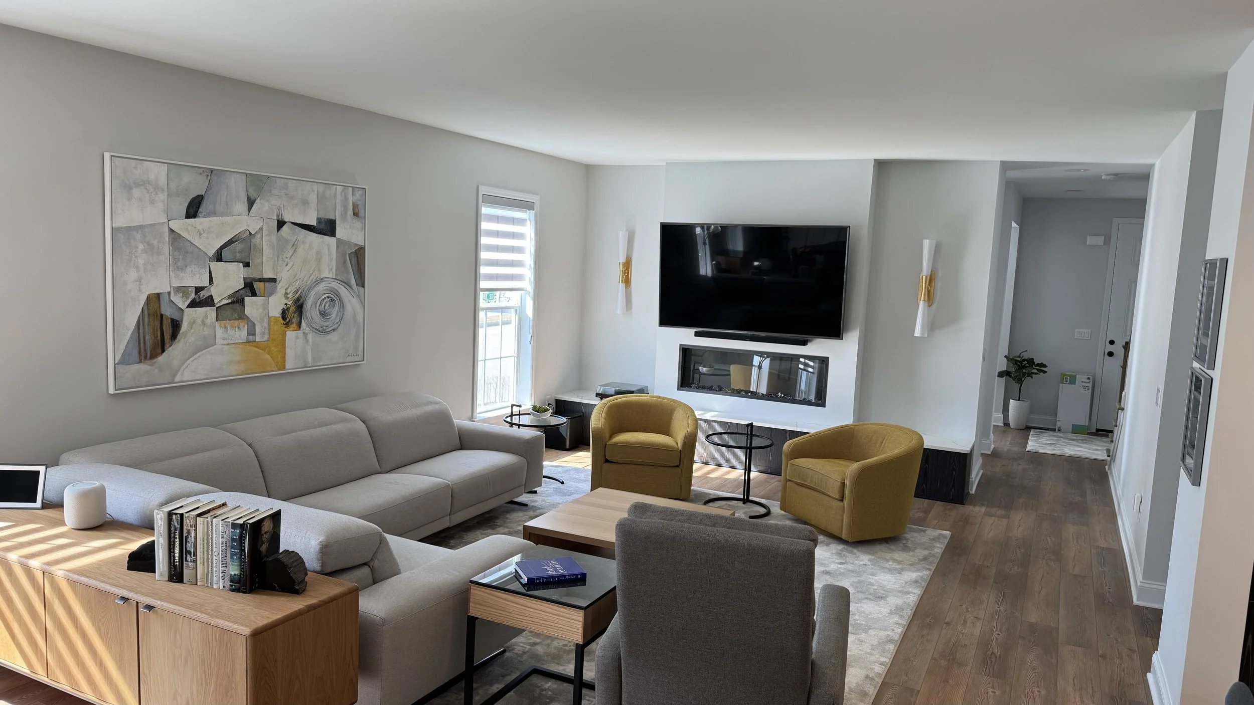 Modern living room with white sectional sofa, yellow armchairs, abstract wall art, wall-mounted TV, fireplace, small tables, potted plant, and hardwood flooring.