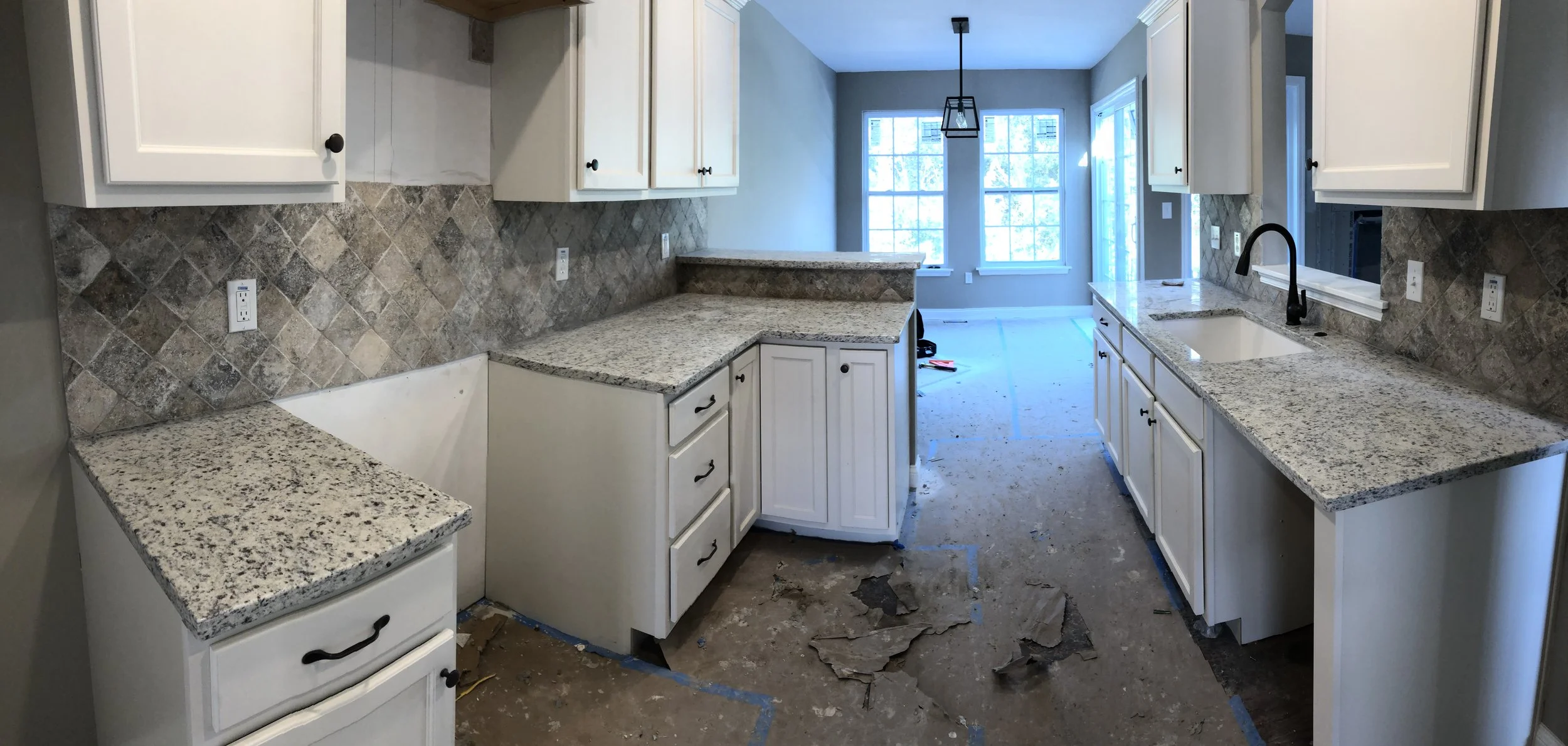 A kitchen under construction featuring white cabinets with black handles, granite countertops, a tiled backsplash, a black sink faucet, and large windows in the background allowing natural light.