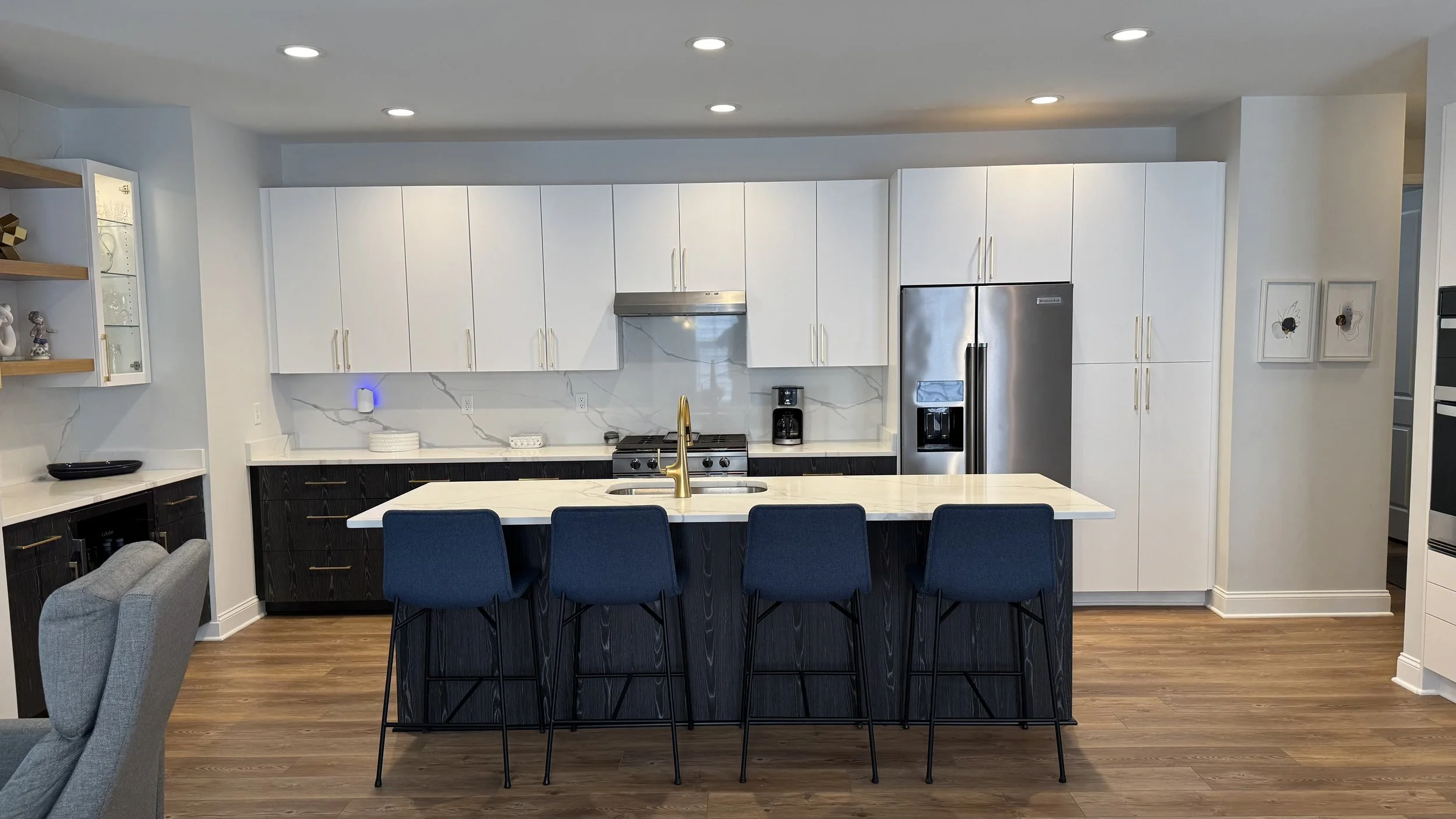 Modern kitchen with white upper cabinets, black lower cabinets, marble backsplash, and a kitchen island with a gold faucet and four blue chairs.
