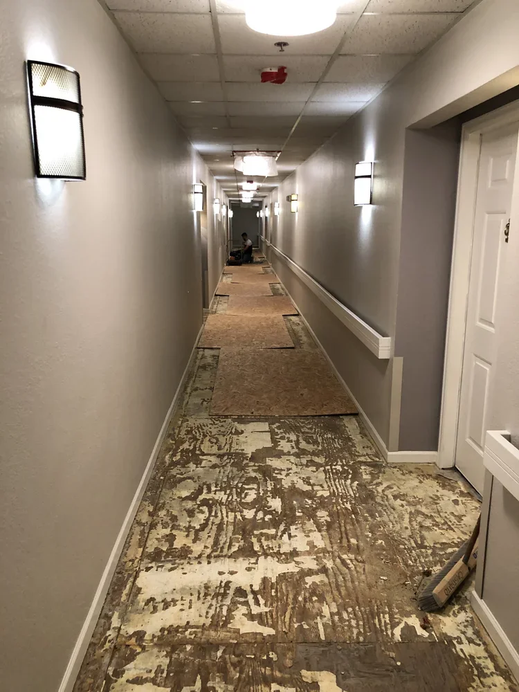 Long hotel corridor with partially removed carpet, beige walls, and wall-mounted lights, with a person sitting at the far end.