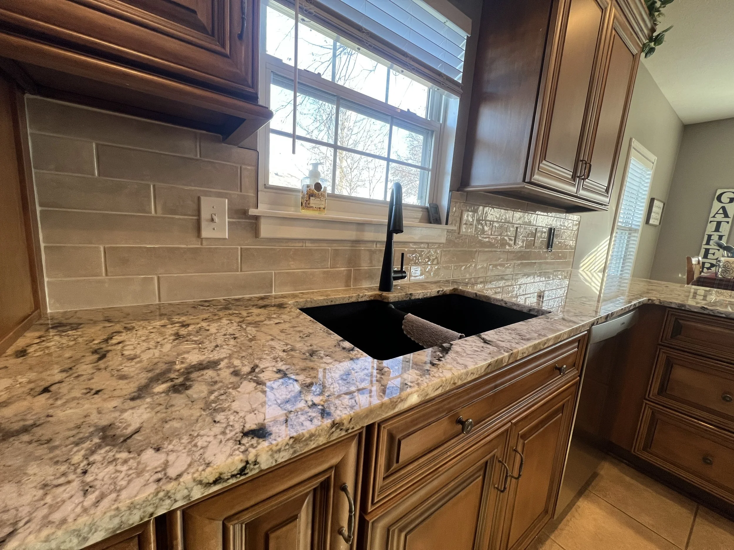 Kitchen countertop with a black sink and faucet, wooden cabinets, a window with blinds, beige tiled backsplash, and granite countertop with brown, beige, and black speckles.