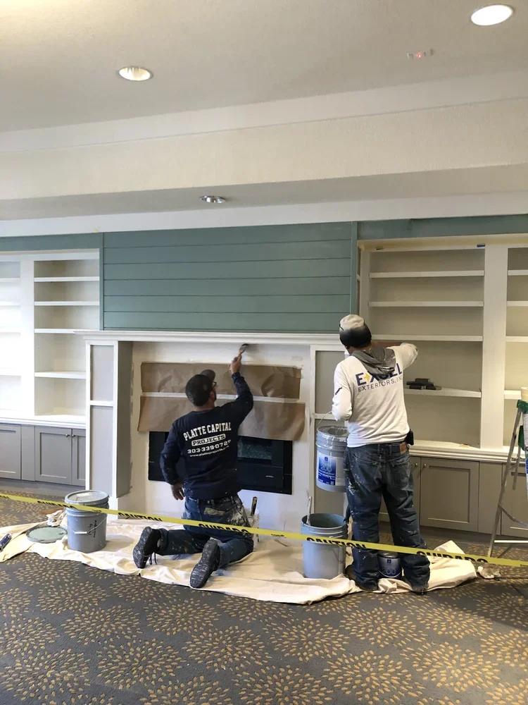 Two workers installing or renovating a fireplace area in a room with built-in shelves, surrounded by construction tools and materials, with caution tape in the foreground.