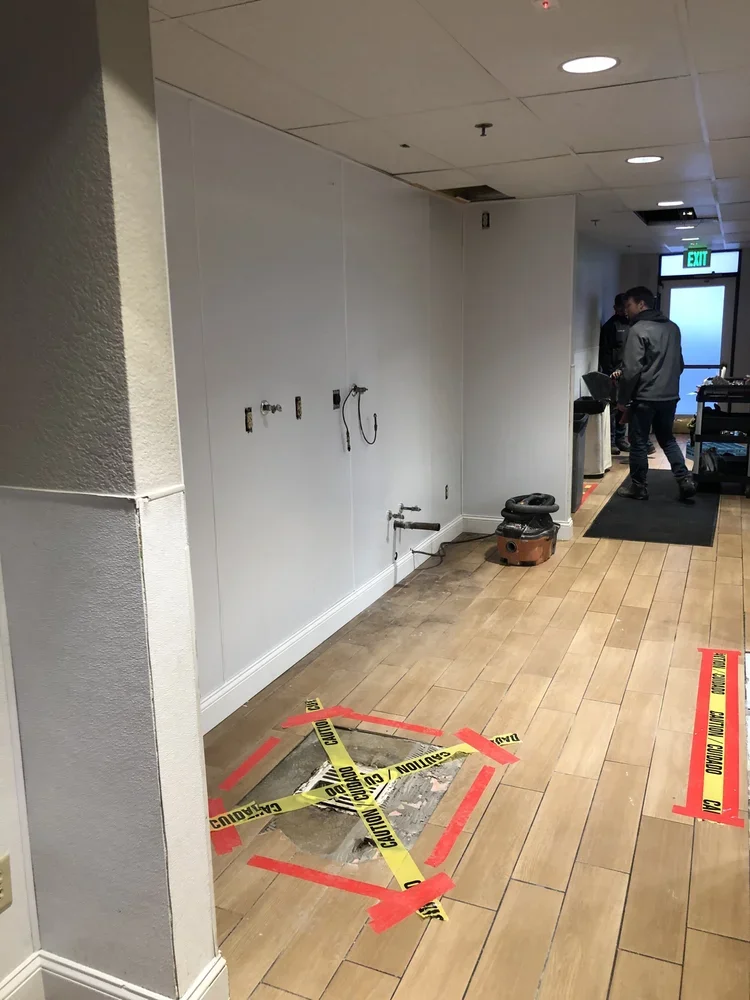 Floor with a hole marked off with caution tape, located in an indoor space with people standing near a doorway.