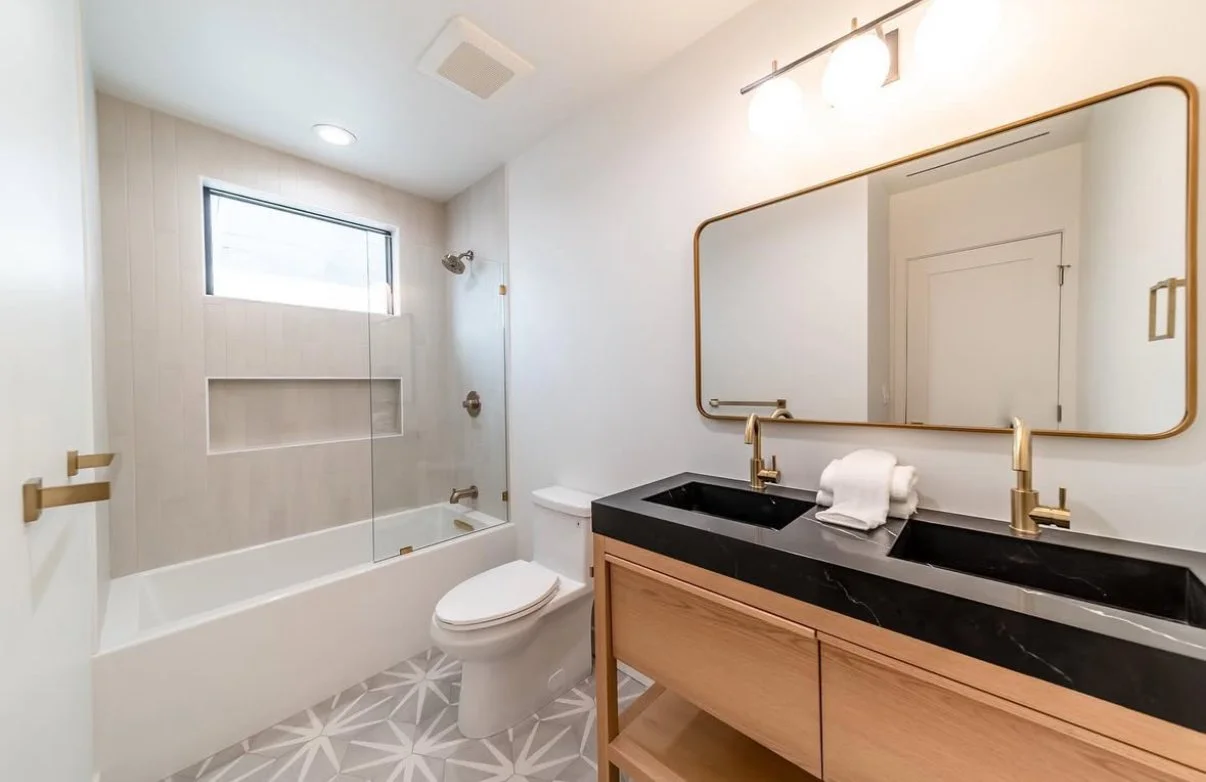 Modern bathroom with a bathtub and shower, a toilet, a wooden vanity with a black countertop and double sinks, a large mirror, and a geometric tile floor.