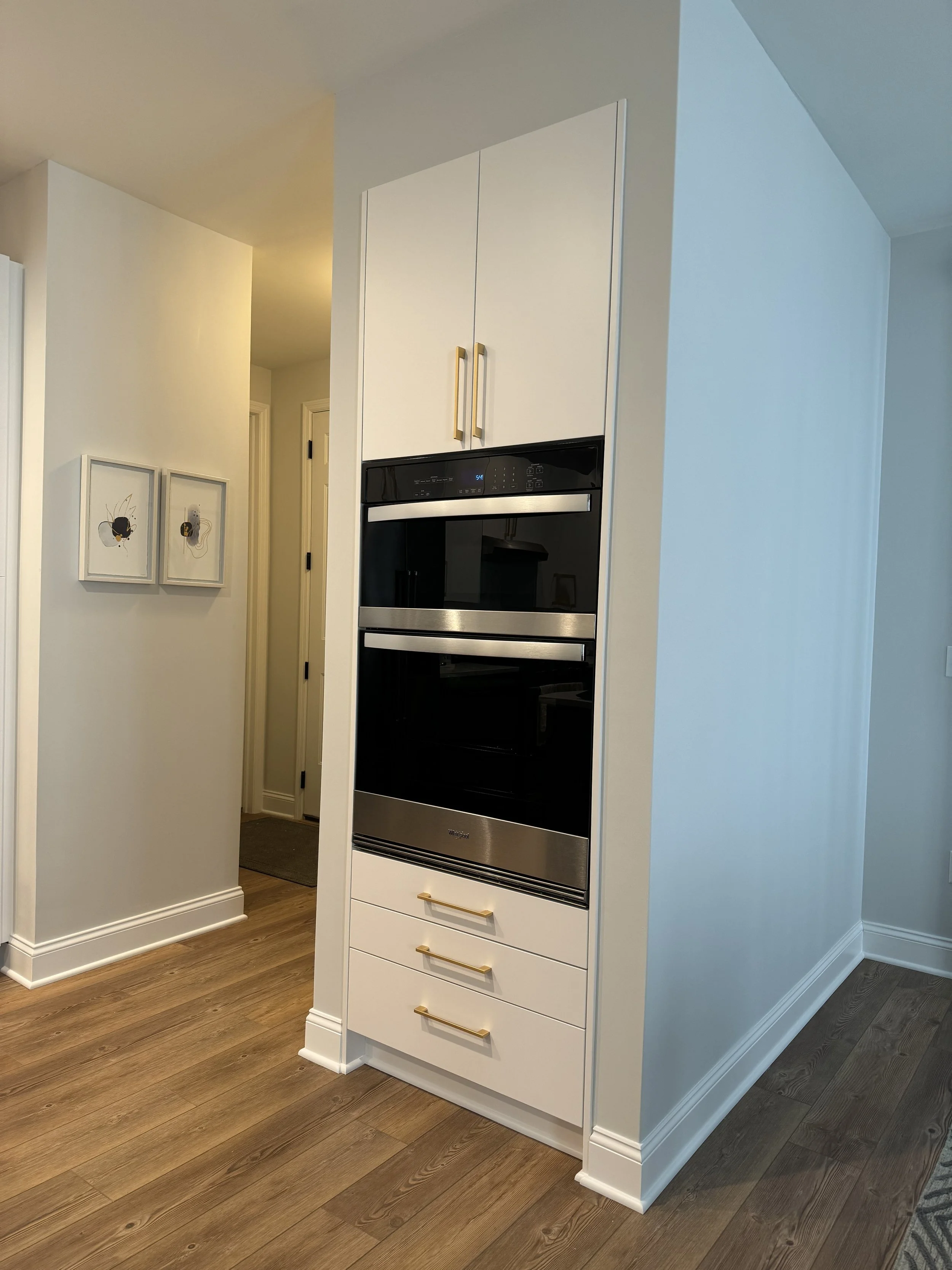 Close-up of a built-in oven integrated into a white kitchen cabinet with drawers, gold handles, and a white wall in the background. There are framed artwork on the wall to the left.