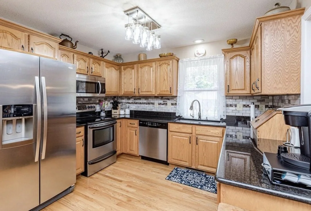 Wooden kitchen cabinets with black countertops, a stainless steel refrigerator, stove, dishwasher, and microwave, with a window above the sink and decorative items on top of cabinets.