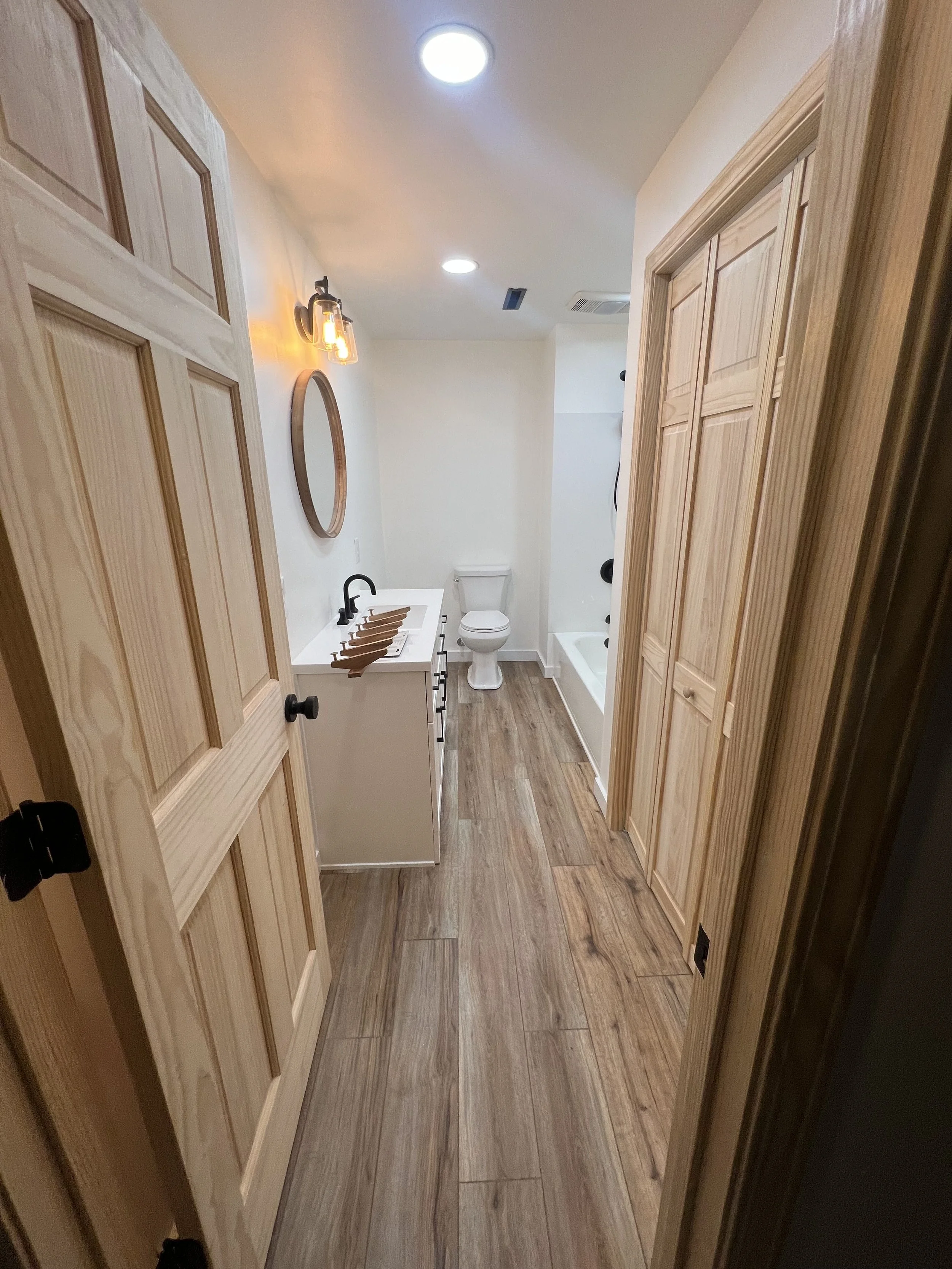 View of a bathroom with wooden doors and flooring, a white vanity with a black faucet, a mirror, a toilet, and a bathtub, with ceiling lights and wall-mounted lighting.