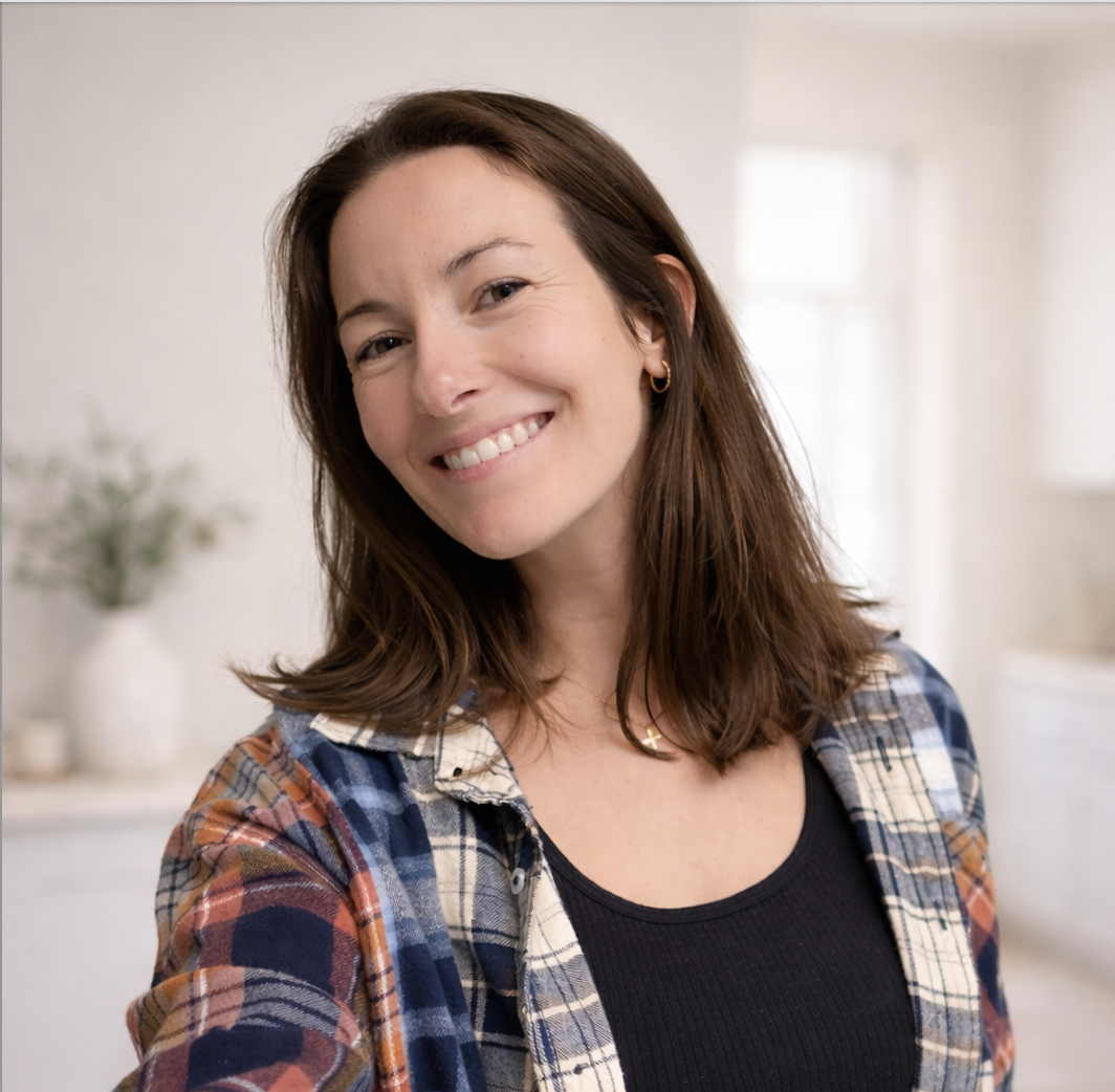A woman with shoulder-length brown hair, smiling, wearing a black top and a plaid shirt, standing indoors in a brightly lit room.