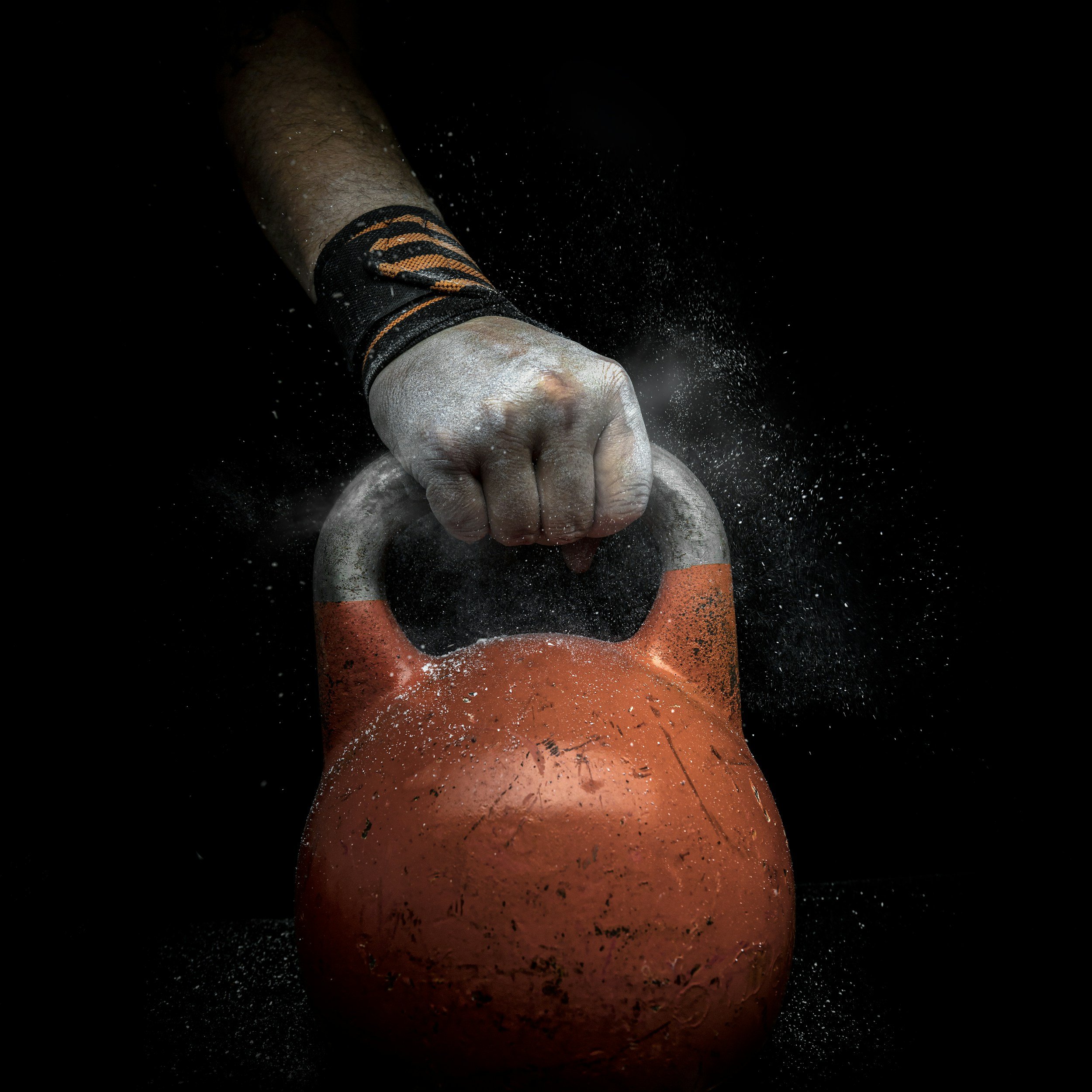 A person's fist with chalk dust, wearing a black and orange wristband, gripping an orange kettlebell against a black background.
