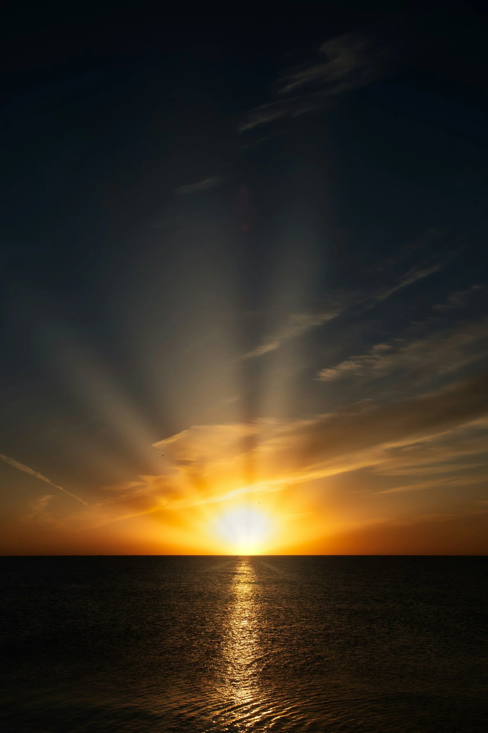 Sunset over the ocean with visible sun rays and partly cloudy sky.