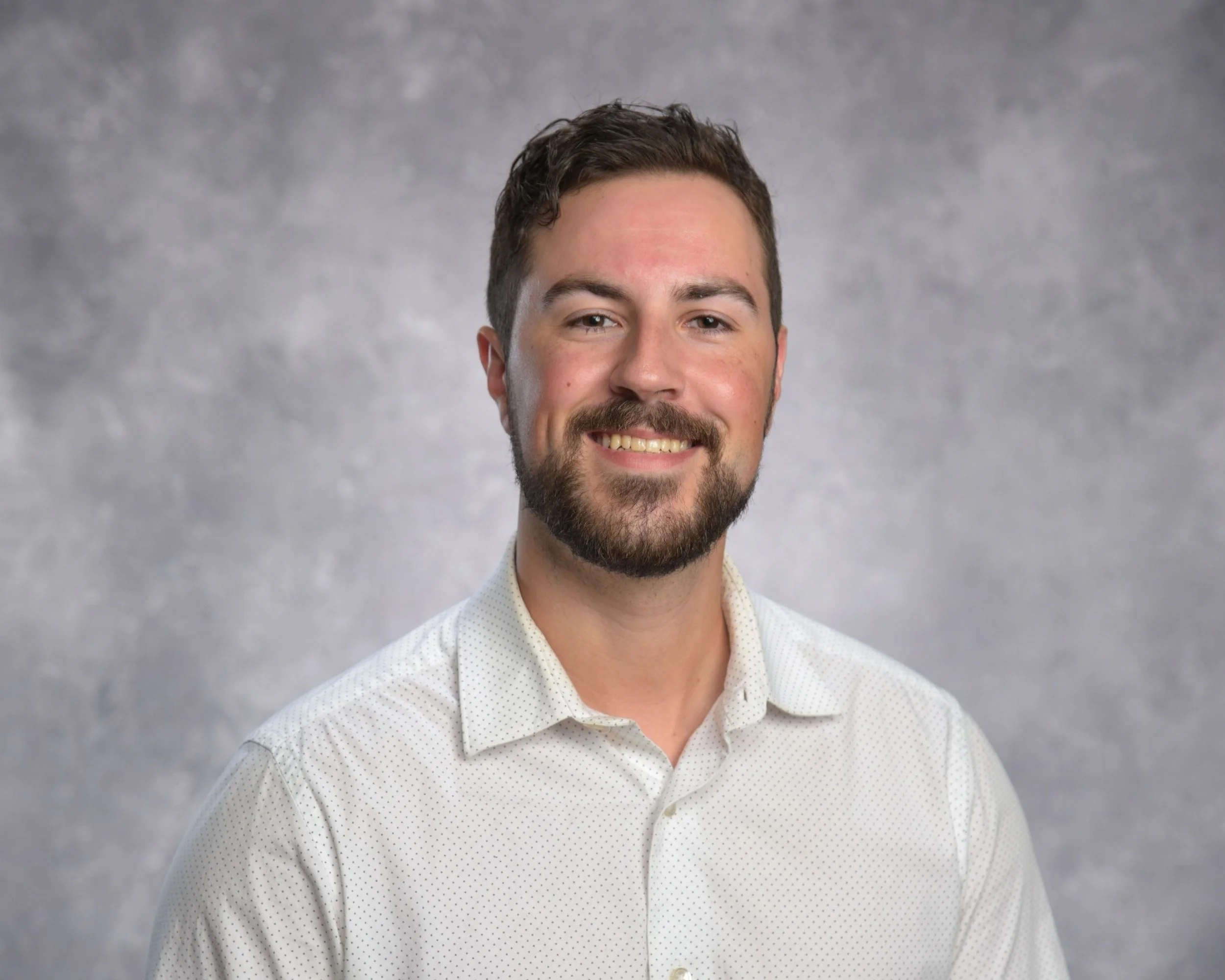 Headshot of a smiling man with short dark hair, beard, and mustache, wearing a white button-up shirt with a subtle pattern, against a gray textured background.
