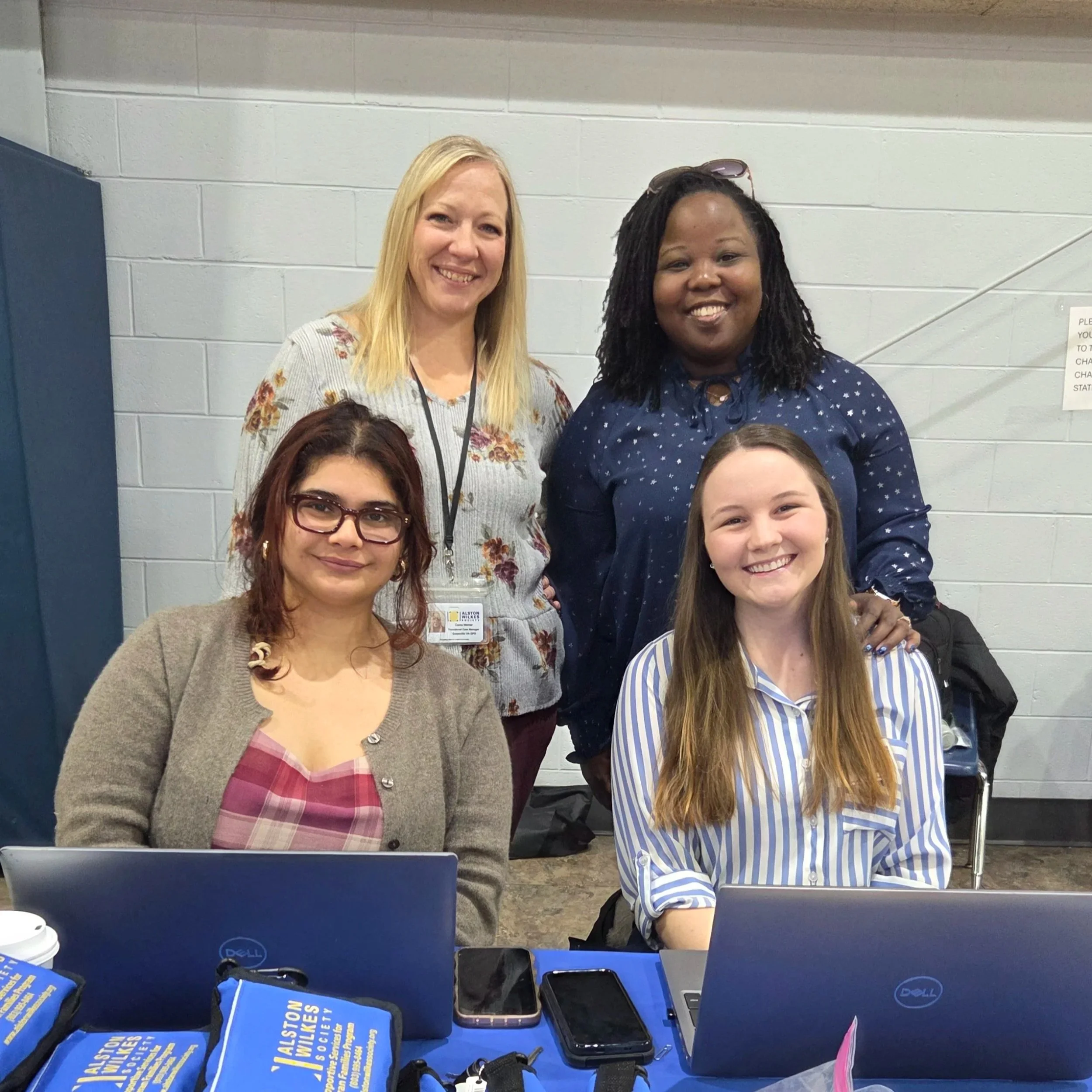 Four women in an indoor setting, two sitting at a table with laptops and two standing behind them, smiling at the camera.
