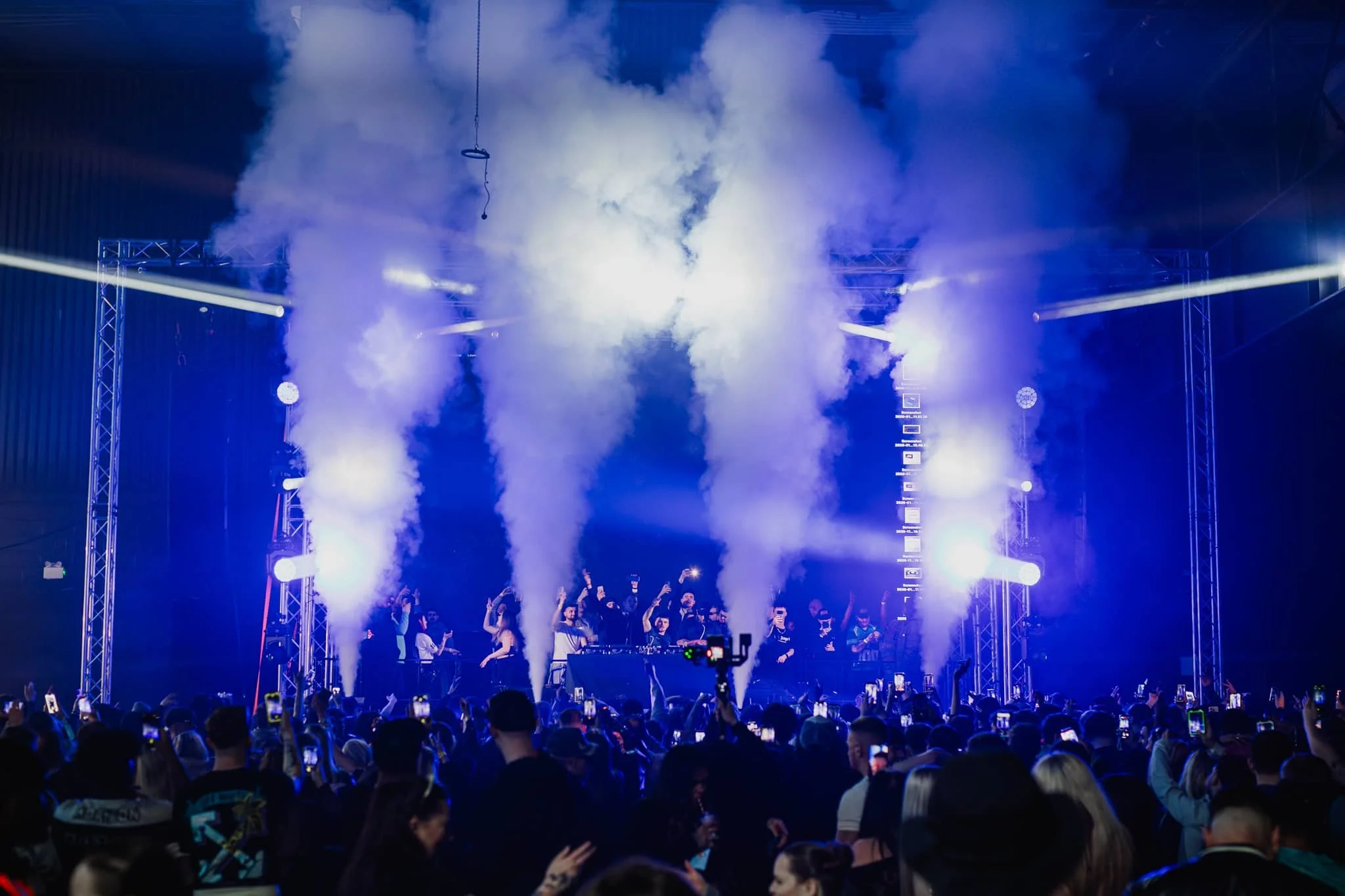 Concert stage with smoke effects, bright stage lighting, and a large crowd of people taking photos and dancing.