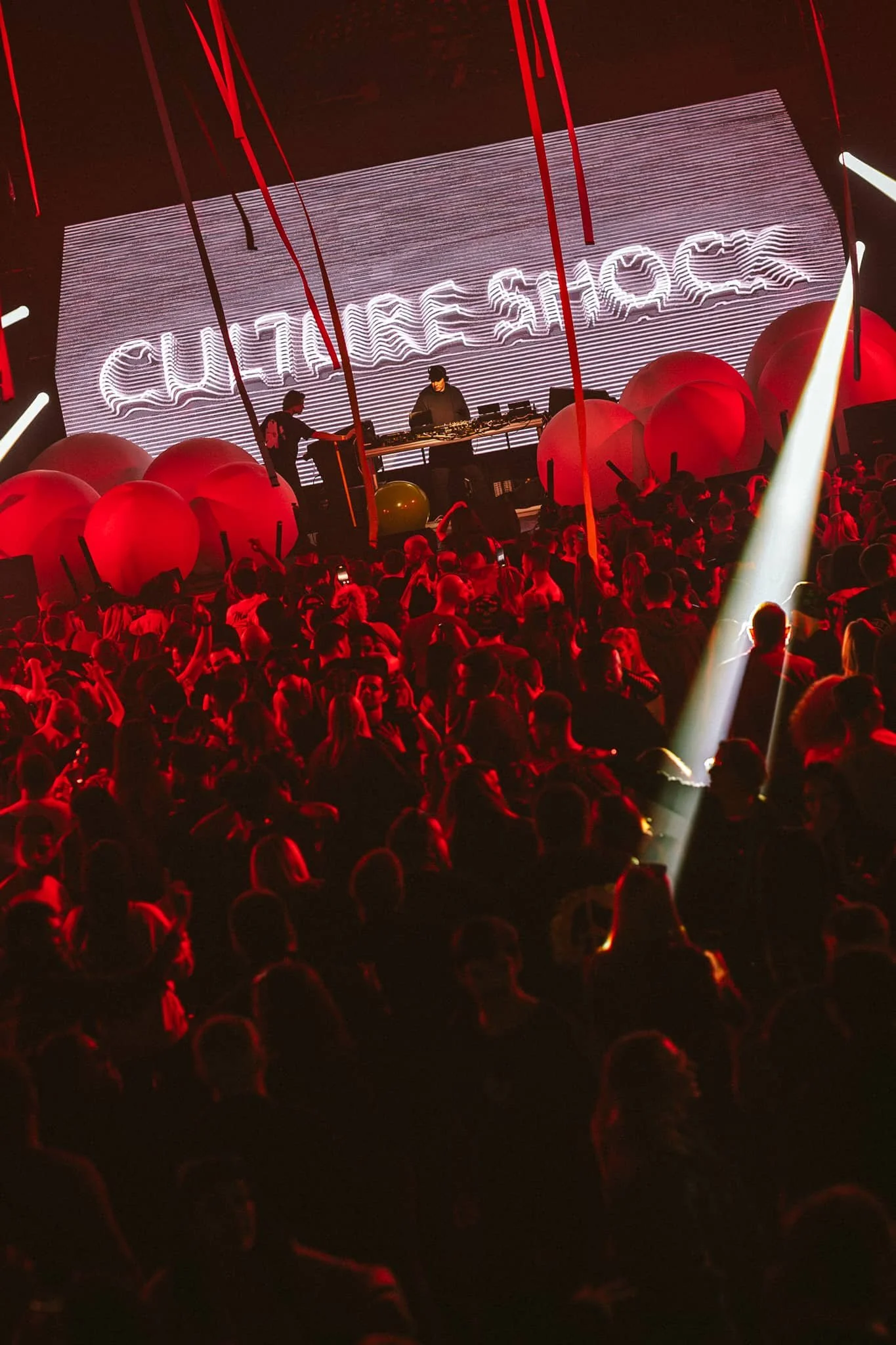 A live DJ performance on stage with a large LED screen displaying the words 'CULTURE SHOCK' behind the DJ, red inflatable balls on stage, and a crowd of people dancing in a dark club with red lighting and a spotlight.