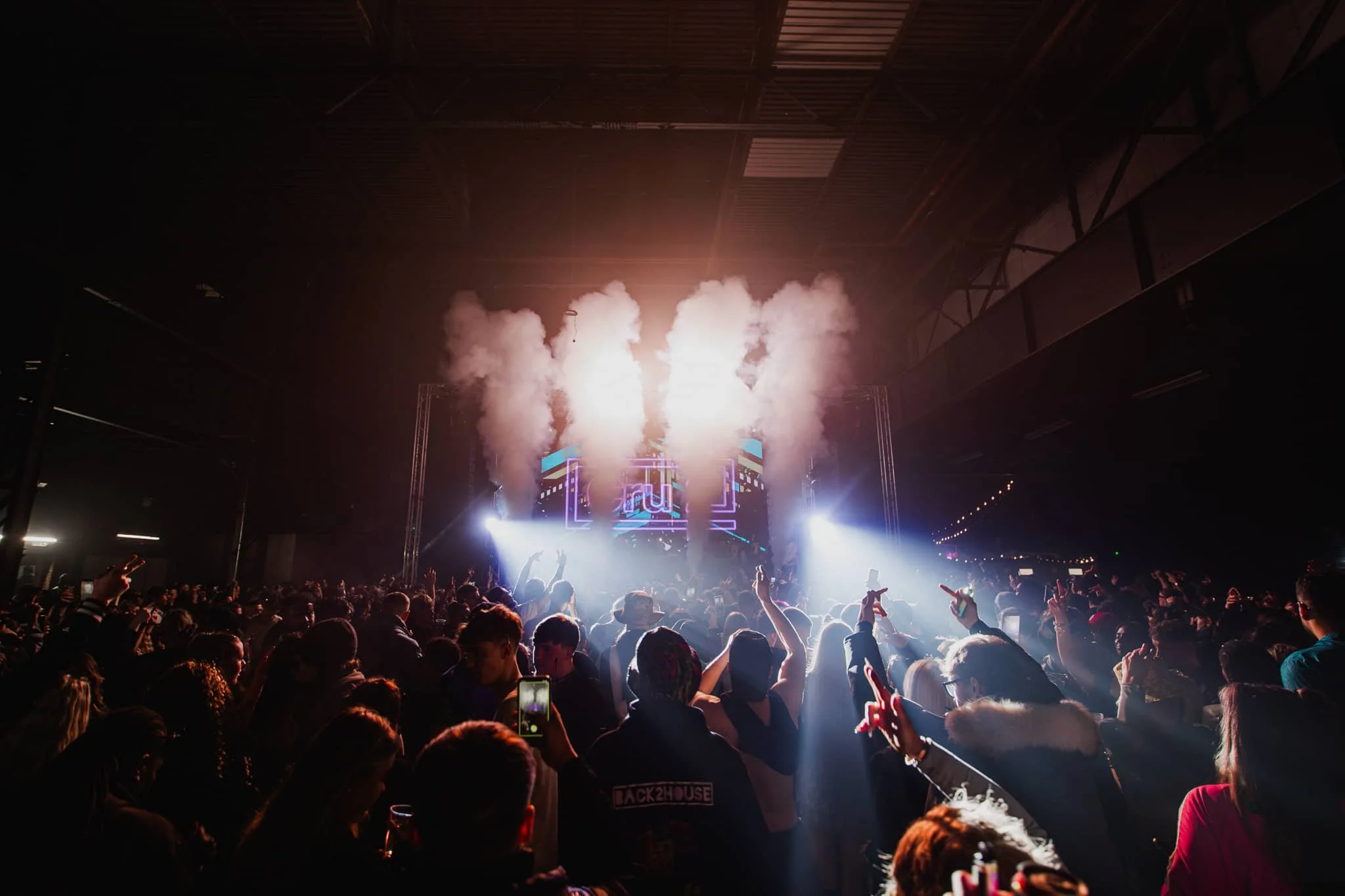 A crowded concert scene with a stage at the front, bright lights, smoke effects, and a DJ or performer. The audience is dancing and taking photos, enjoying the music in an indoor venue.