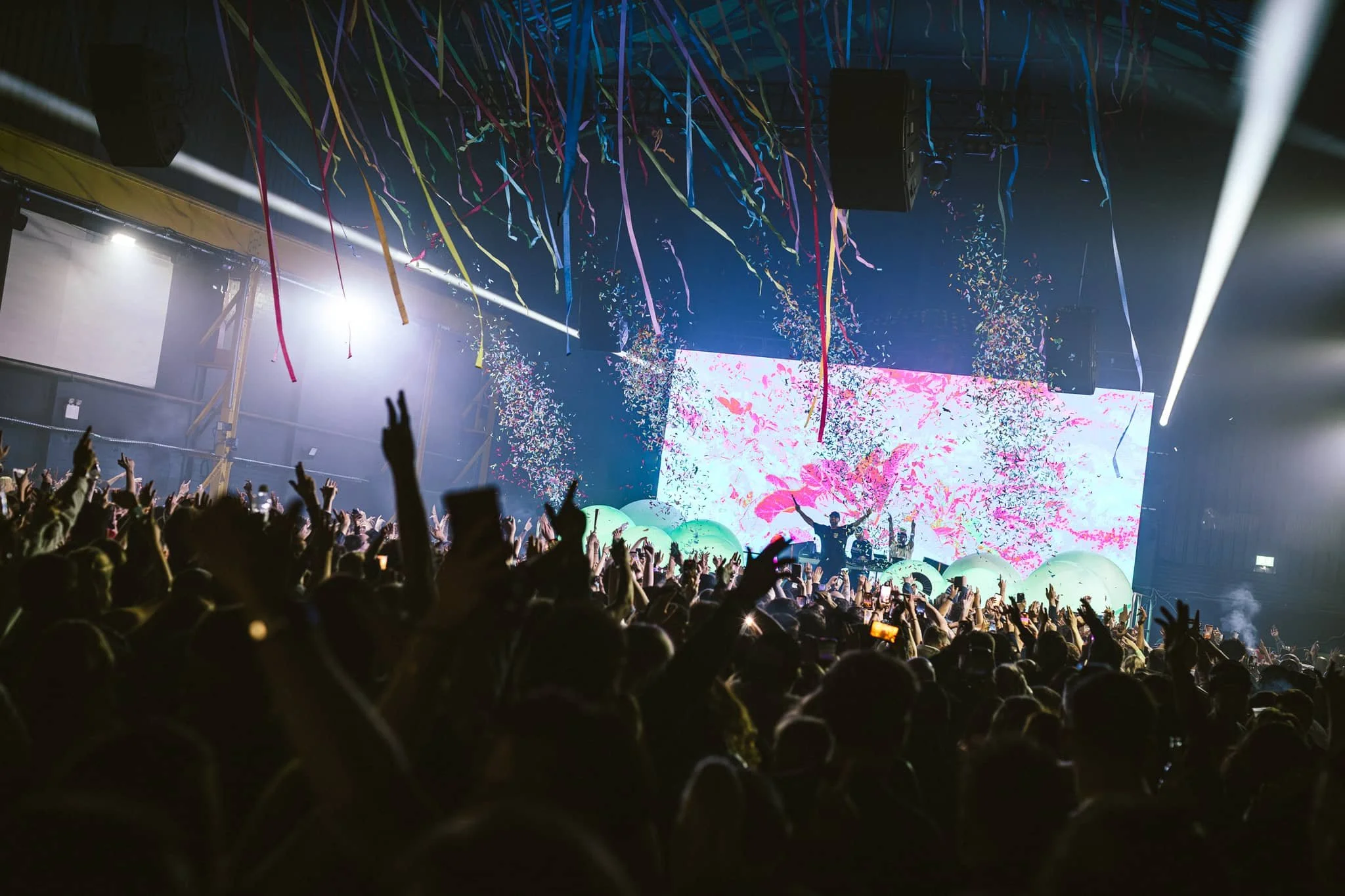 Crowd of people at a concert or festival with arms raised, colorful streamers and confetti falling, and a bright digital screen displaying vibrant pink and white abstract visuals on stage.