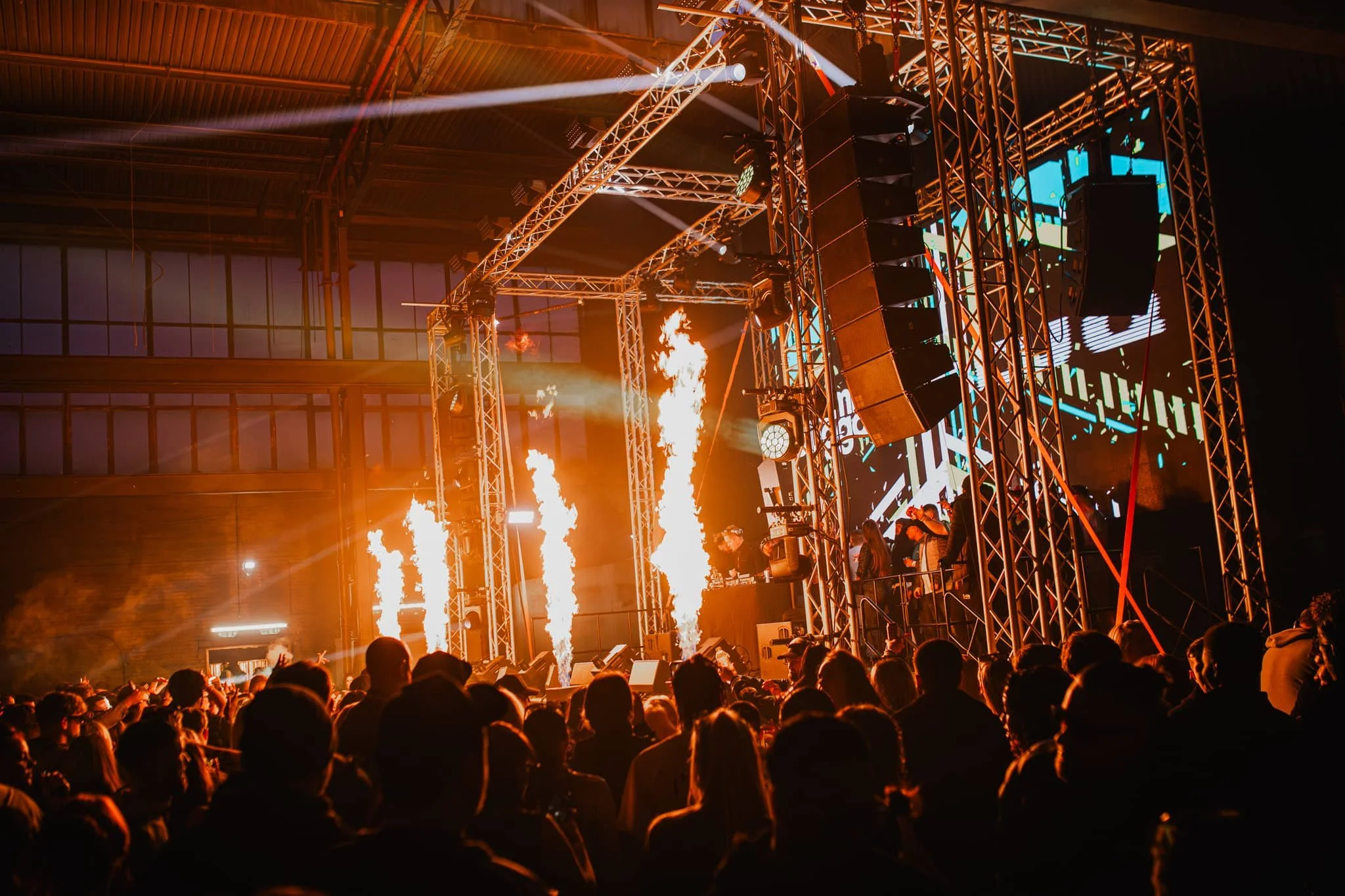 A large crowd watching a concert with pyrotechnics and bright lights on a stage with a digital screen backdrop.
