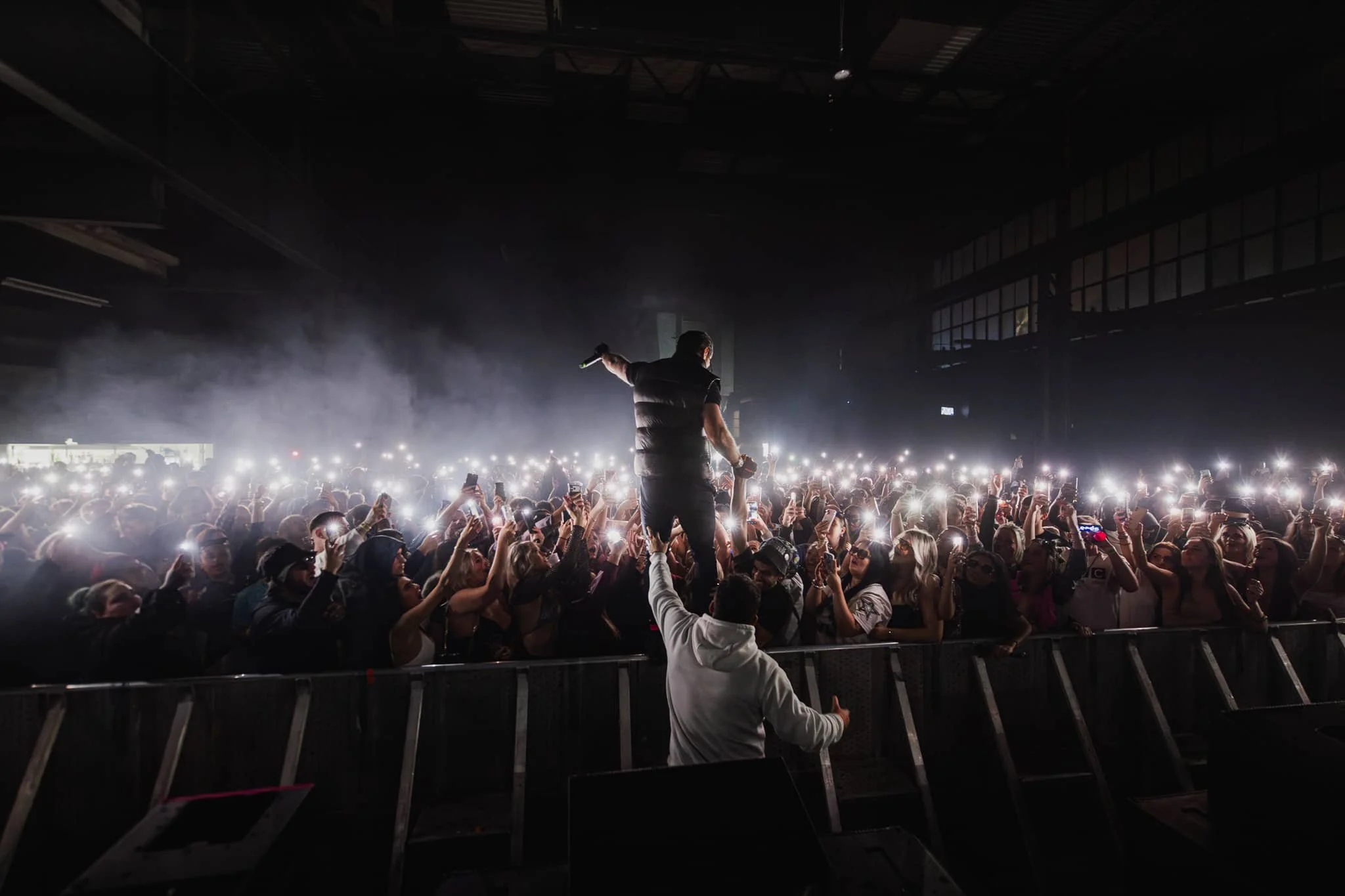 A performer holding a microphone and standing on the stage amidst a crowd at a concert, with many audience members recording and taking pictures with their phones.