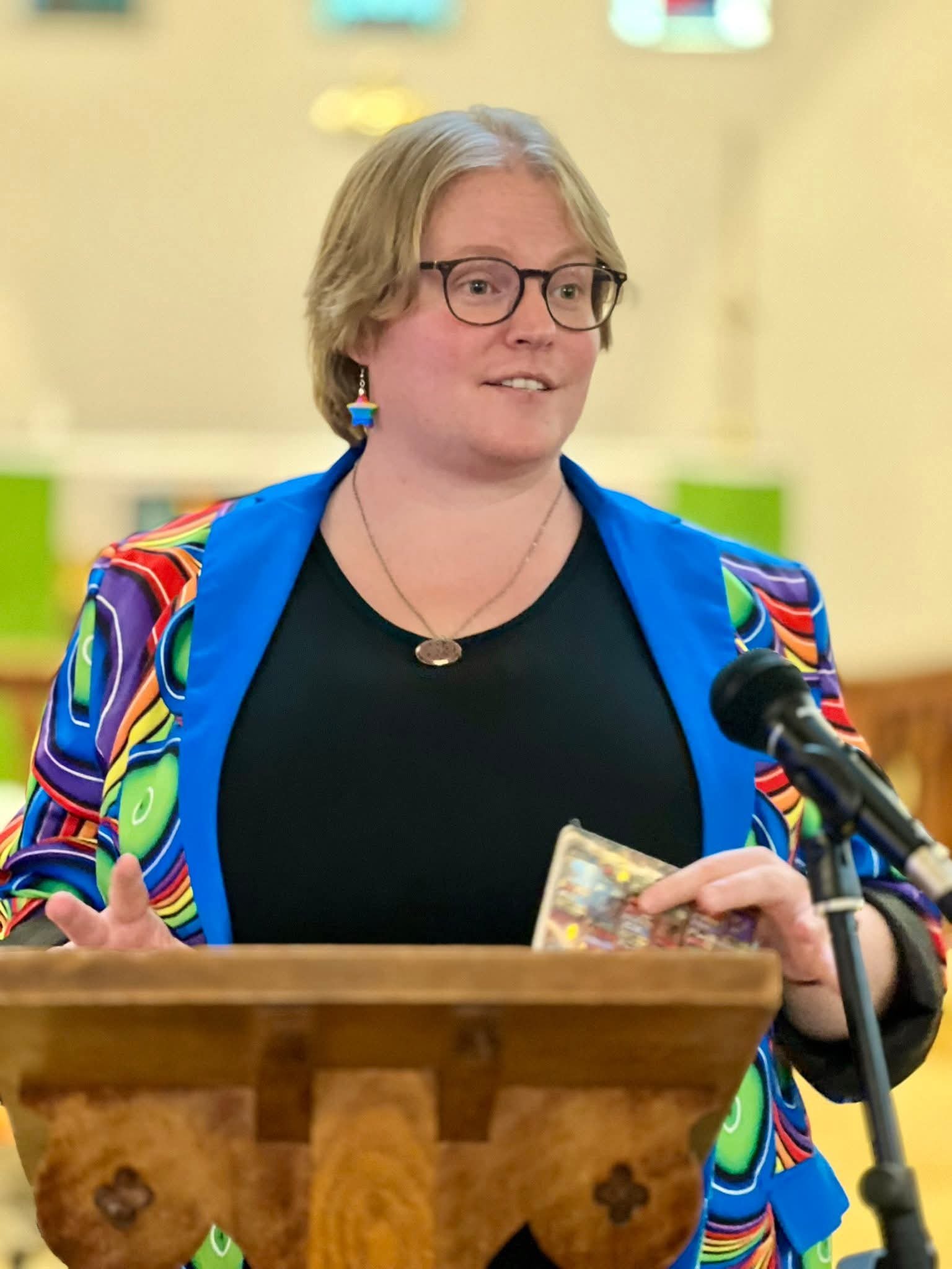 A person with short ginger hair, glasses, and colorful earrings standing at a podium and speaking into a microphone, wearing a vibrant, multicolored jacket and black shirt.