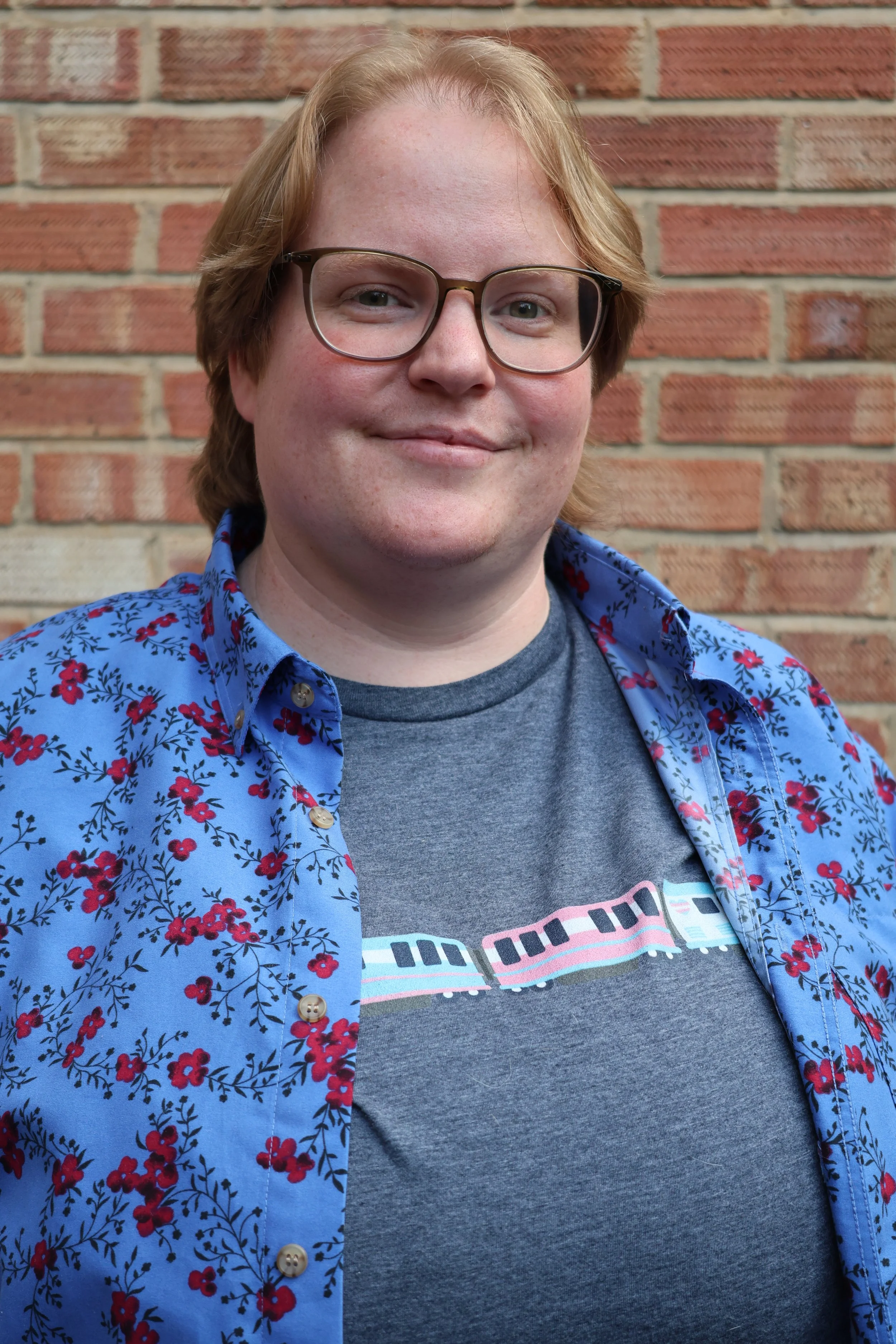 A person with light skin, glasses, and shoulder-length light ginger hair smiling, wearing a blue floral button-up shirt over a gray T-shirt with a train graphic, standing in front of a brick wall.