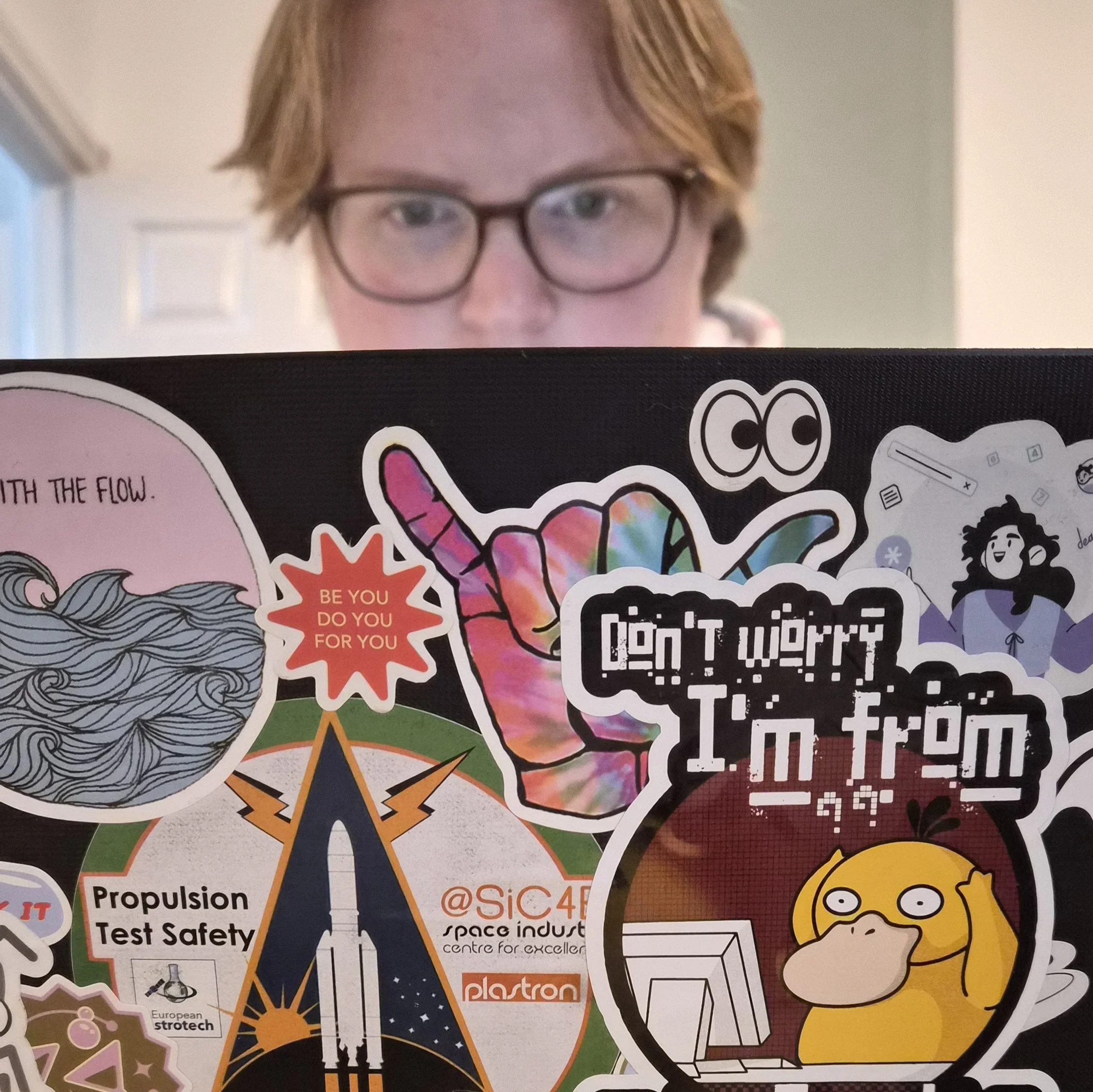 Person with in glasses partially visible behind a laptop covered with various colorful stickers, including a cartoon hand making a shaka sign, a yellow creature at a computer, and various text and images.