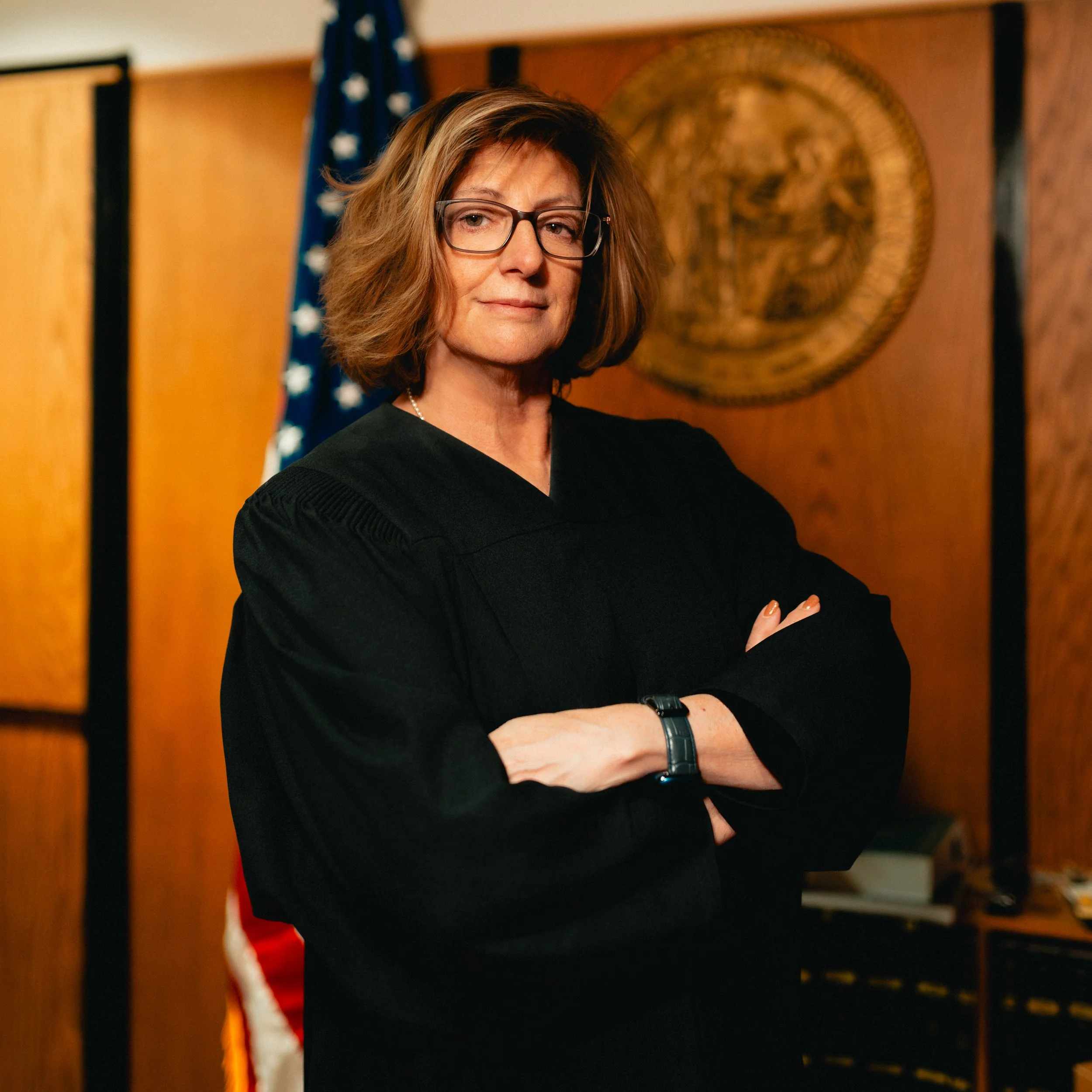 A woman in a black judicial robe stands with her arms crossed in a courtroom, with an American flag and a wooden emblem in the background.