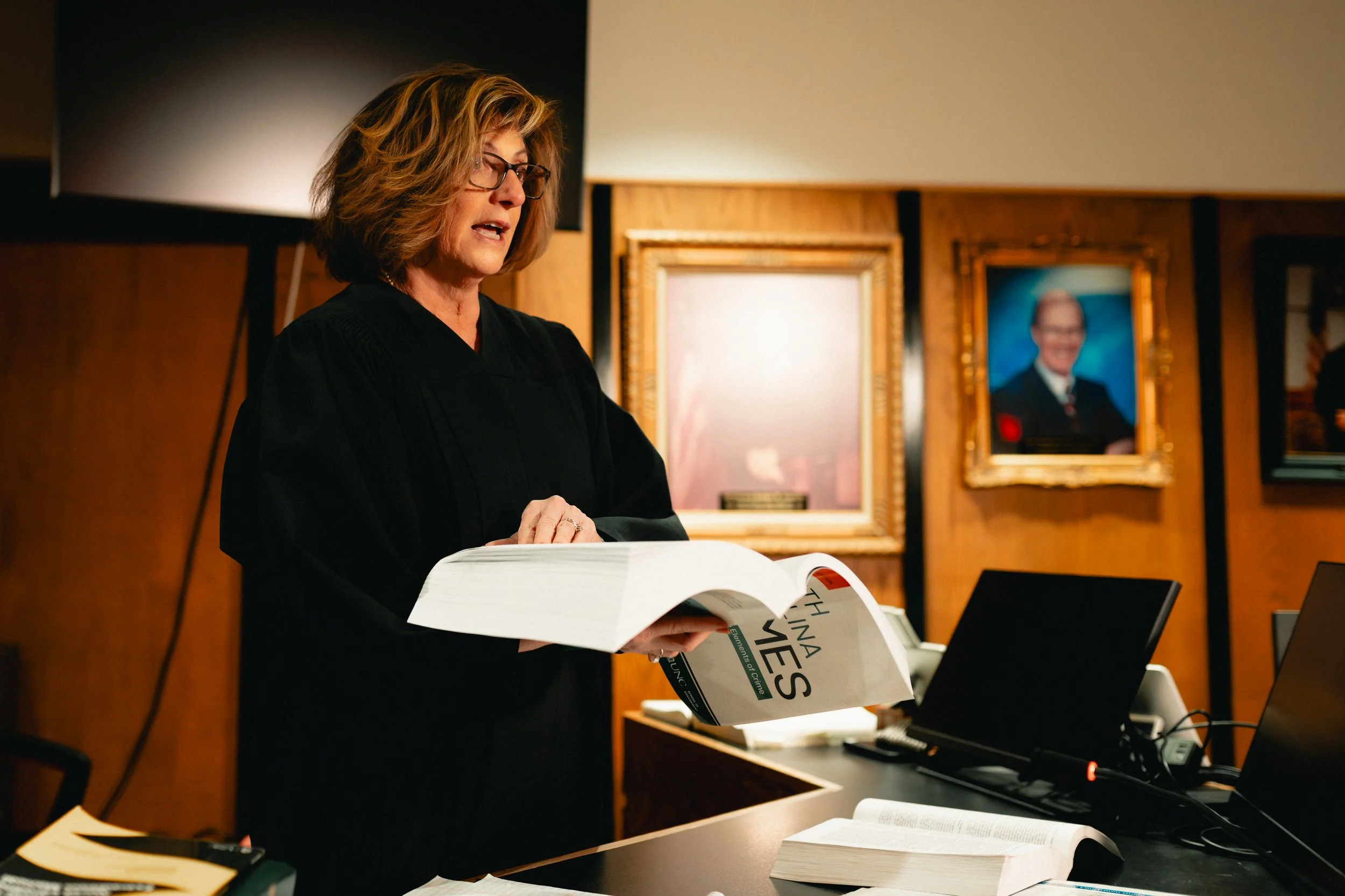 A woman in a black robe, likely a judge, standing in a courtroom reading from a large book. In the background, there are framed portraits, including a man in a suit and glasses.
