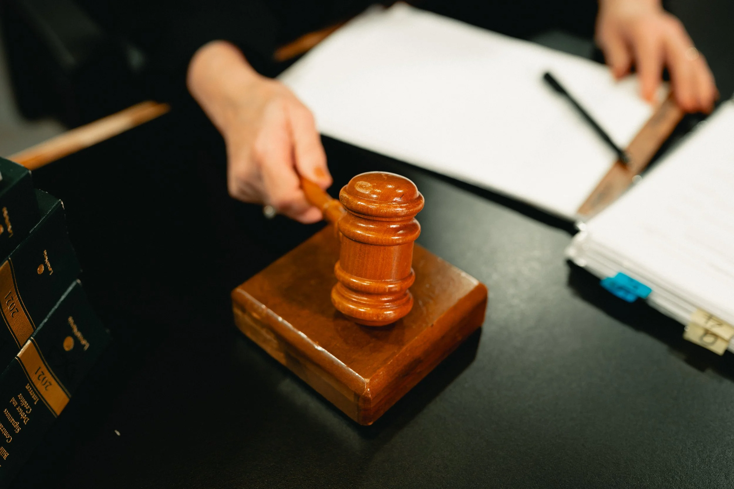 A judge’s wooden gavel on a sound block, with a person holding a gavel and a clipboard with papers and a pen nearby on a desk.
