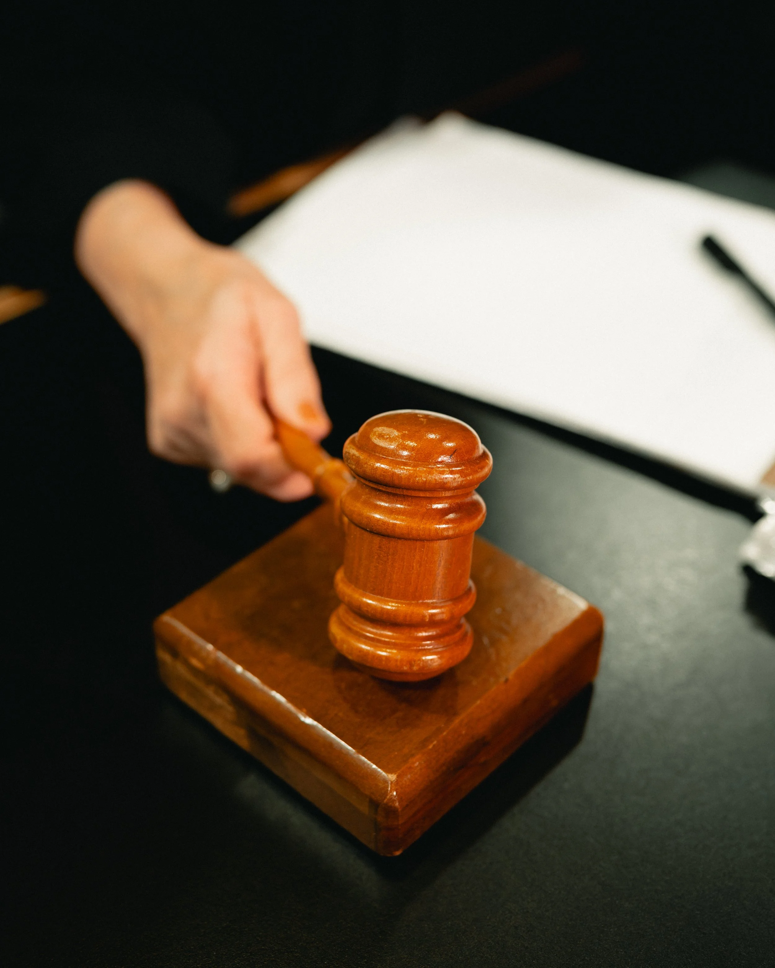 A person holding a wooden gavel over a black table, with a white notepad and pen in the background.
