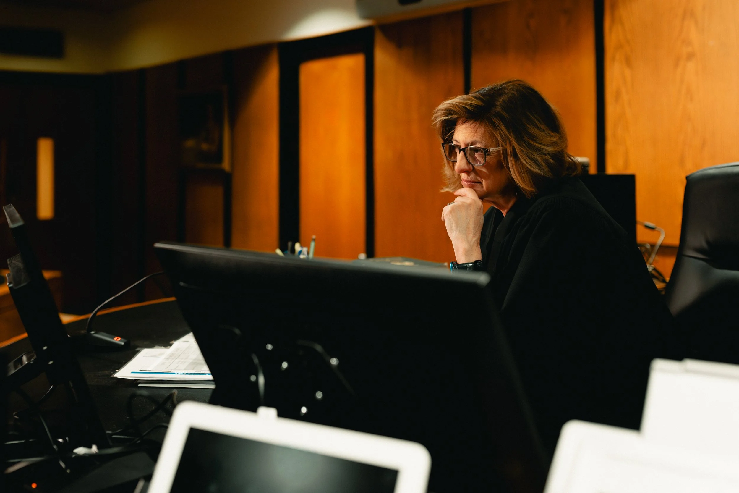 A woman with glasses and shoulder-length hair in a courtroom, seated at a table with a computer monitor and documents, deep in thought, with a wood-paneled wall in the background.