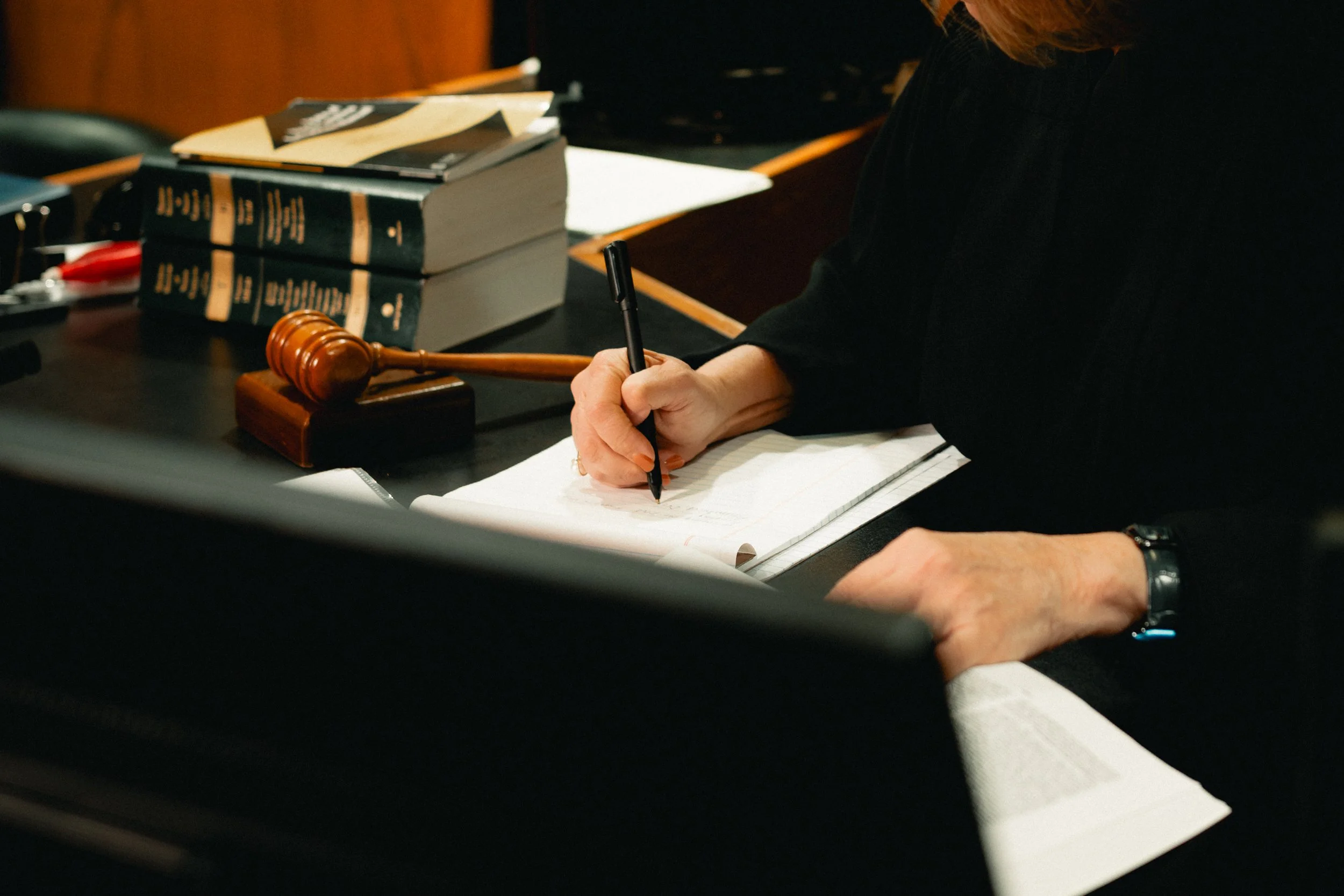 A judge's gavel, books, and legal documents on a desk during a court session, with a person in black robes writing notes.