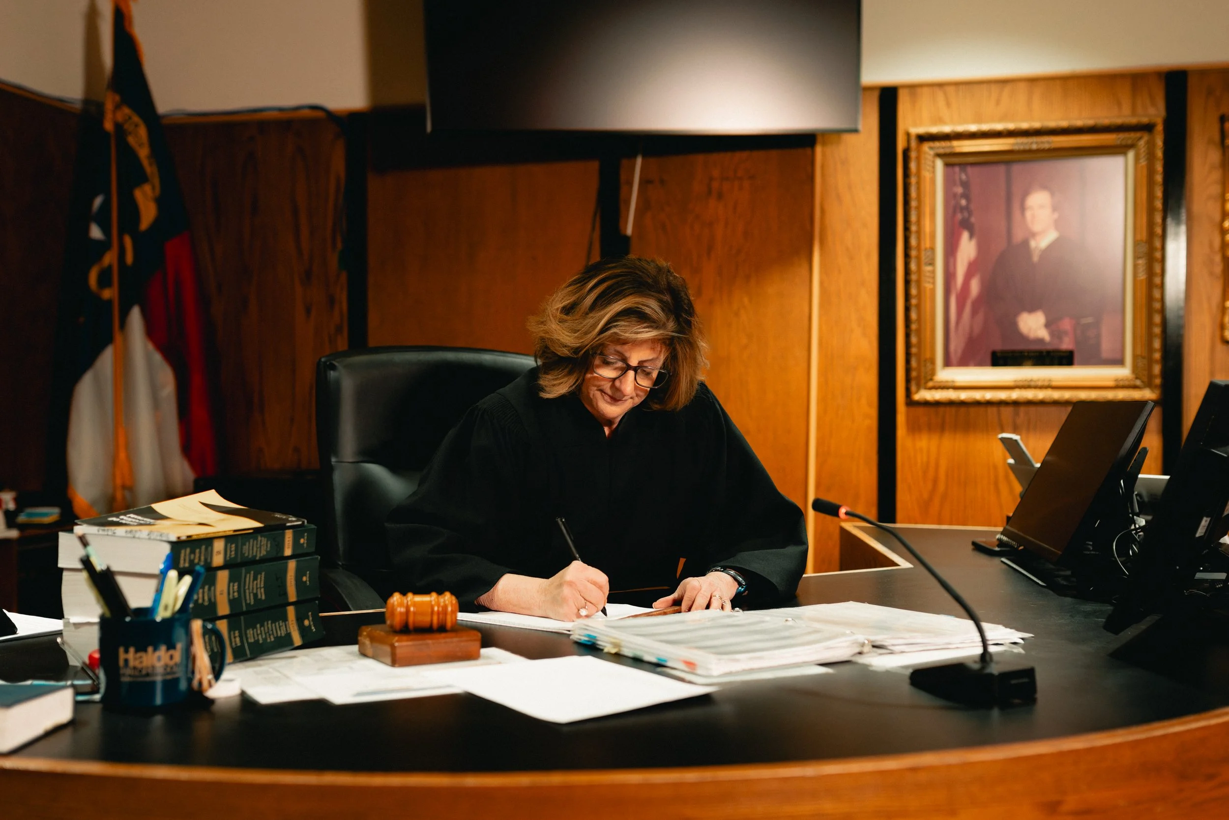 A female judge sitting at a wooden desk, writing on documents, in a courtroom with legal books, a gavel, and framed portrait on the wall.